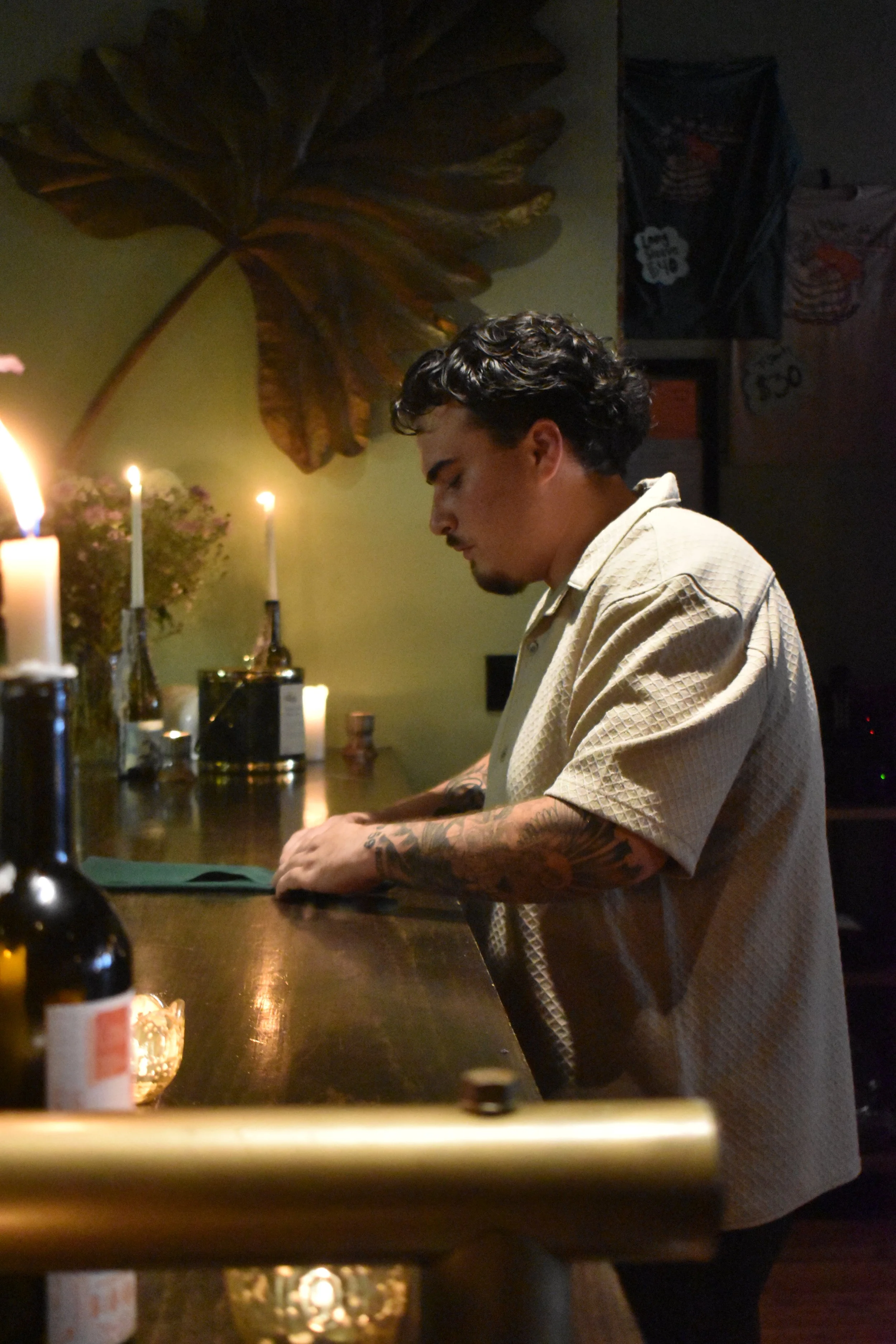 A young man with colored tattoos on his arms and styled black curly hair is sitting at a bar with candles and bottles in the background, with eyes closed and head bowed.