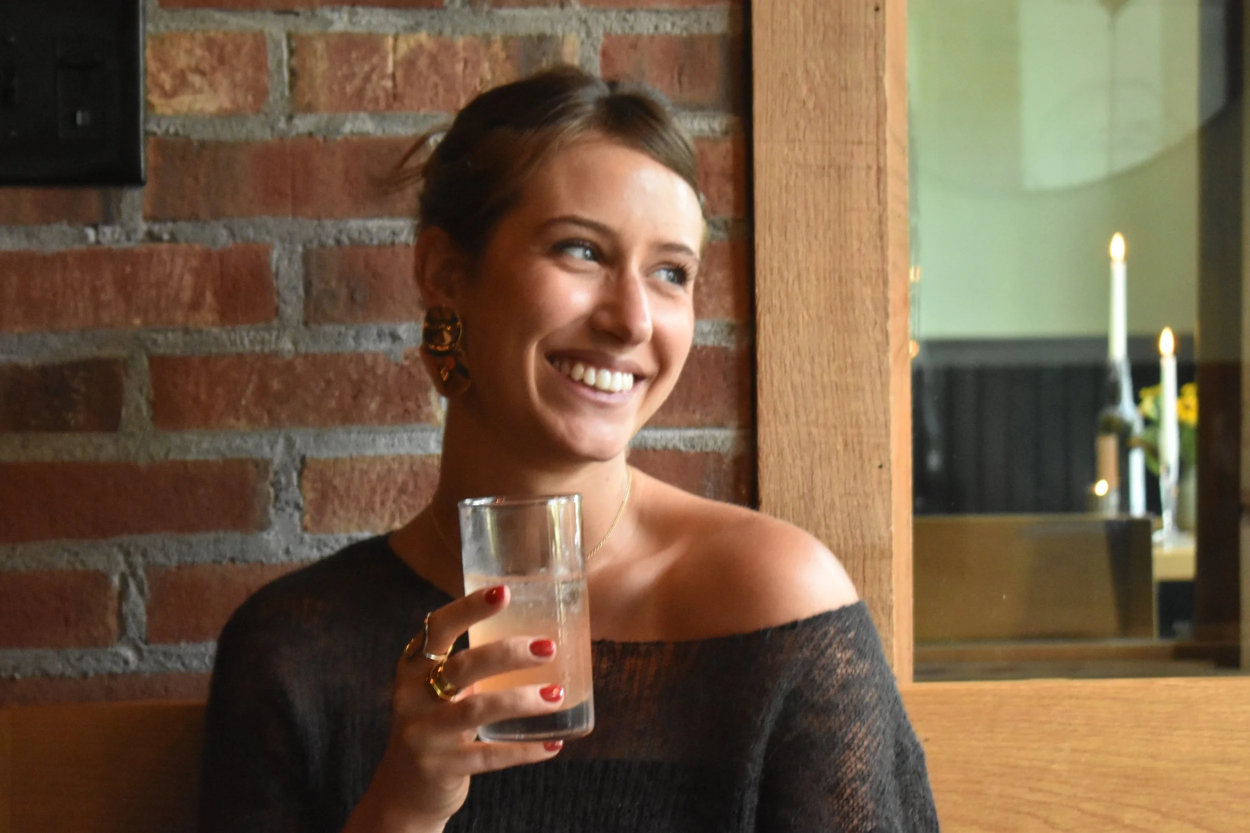 A woman smiling and holding a glass of peach spritz in a restaurant with brick wall background and candles in the distance.