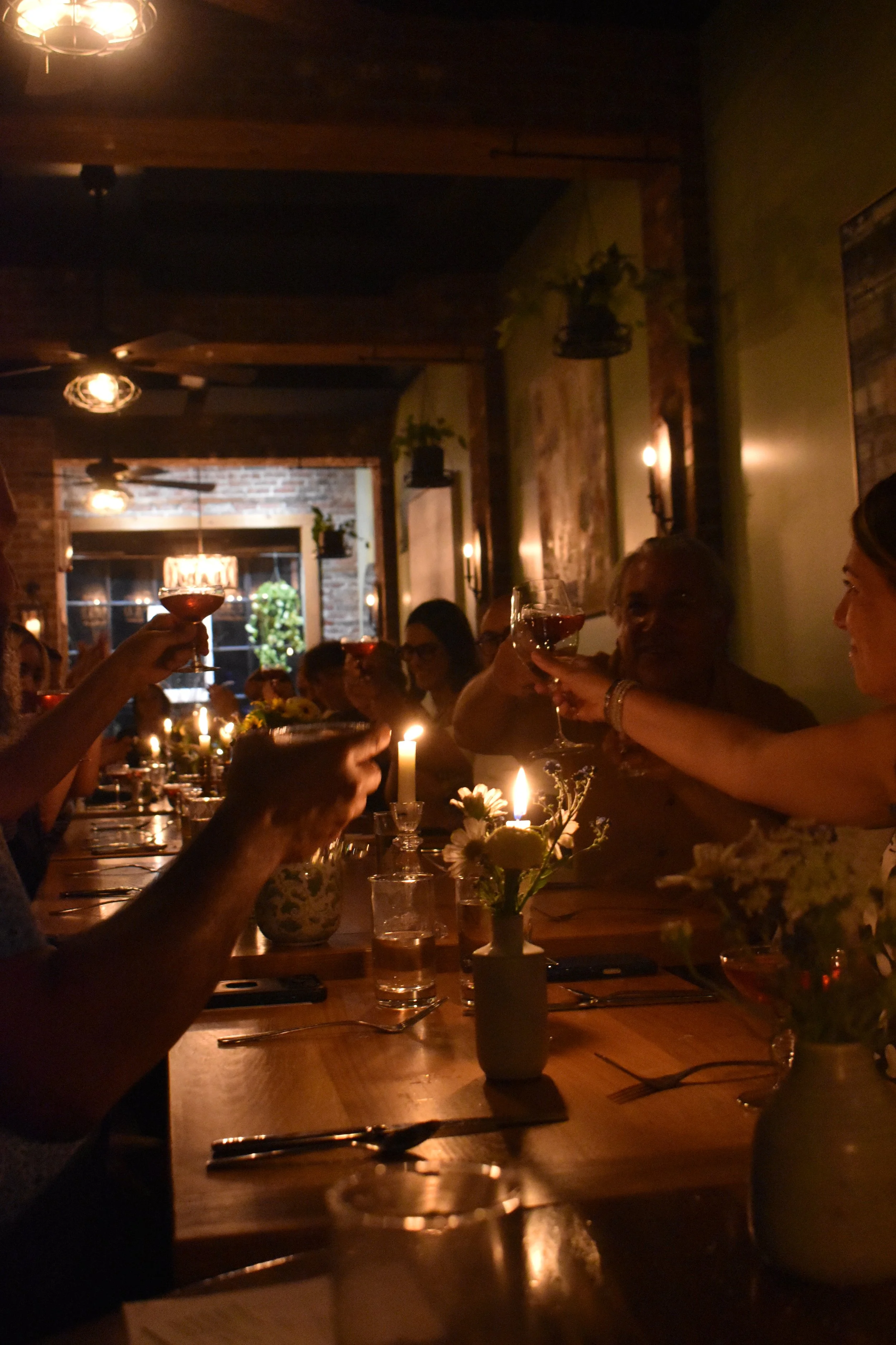 A dimly lit restaurant with people raising glasses in a toast, decorated with flowers and candles on the table.