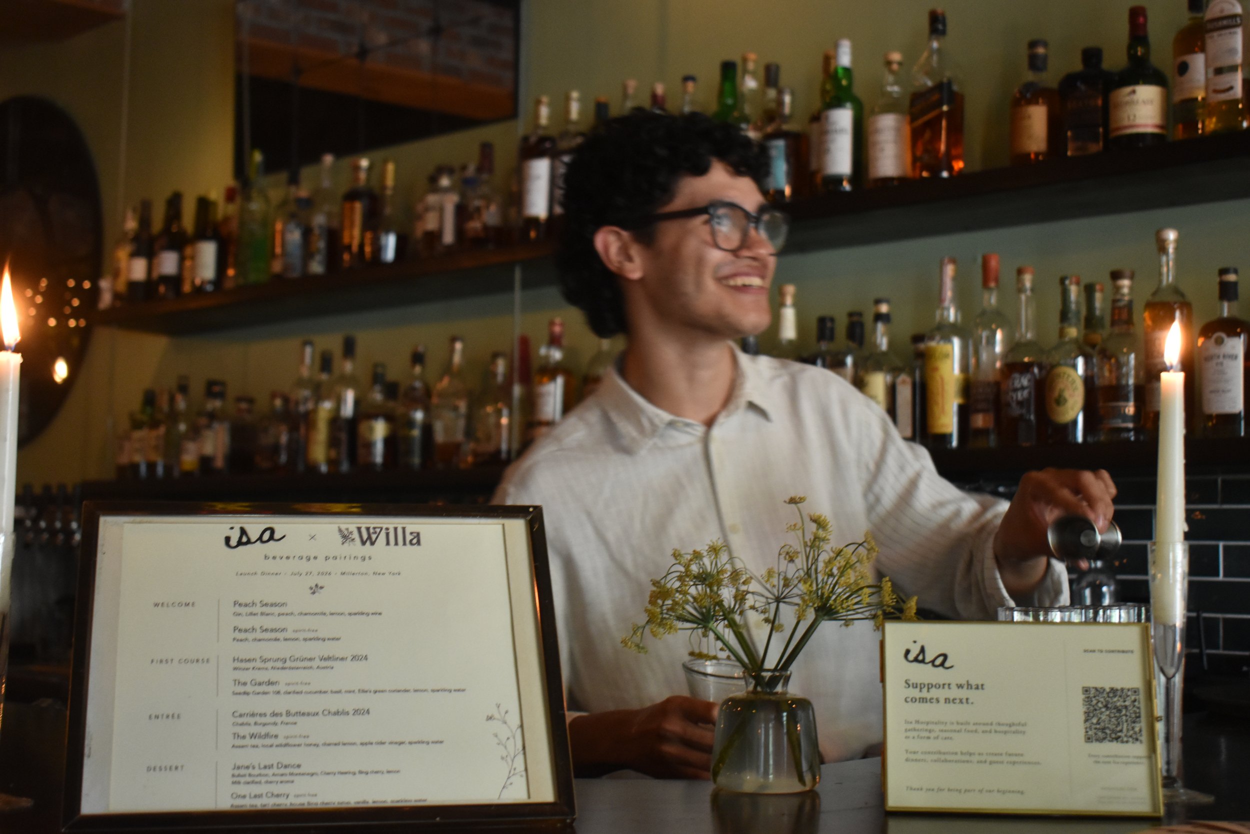 A smiling man wearing glasses and a white shirt behind a bar with an extensive collection of liquor bottles on shelves. There are candles and a menu display on the counter in the foreground, and a green wall in the background.