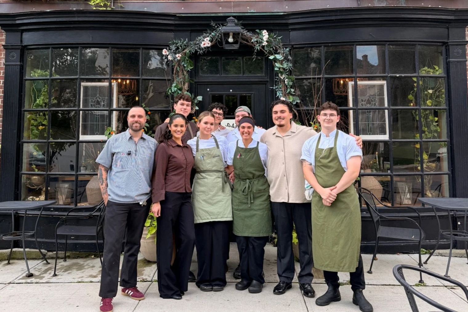 Group of nine people, including four women and five men, standing outside a restaurant with large windows and decorative floral arch. Some are wearing aprons, indicating they are staff members.