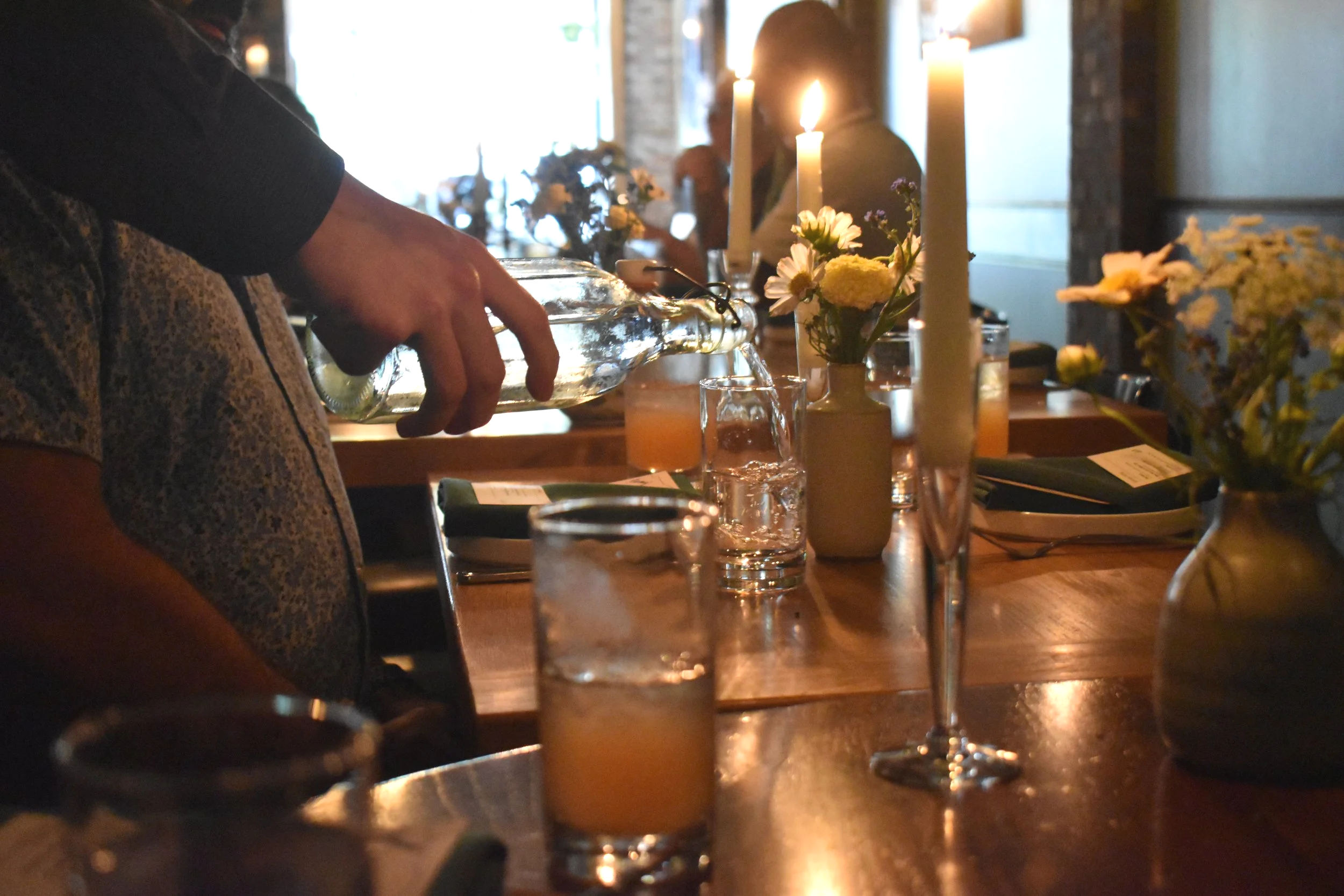 A person pouring a drink at a dimly lit restaurant or bar table decorated with candles and flowers.