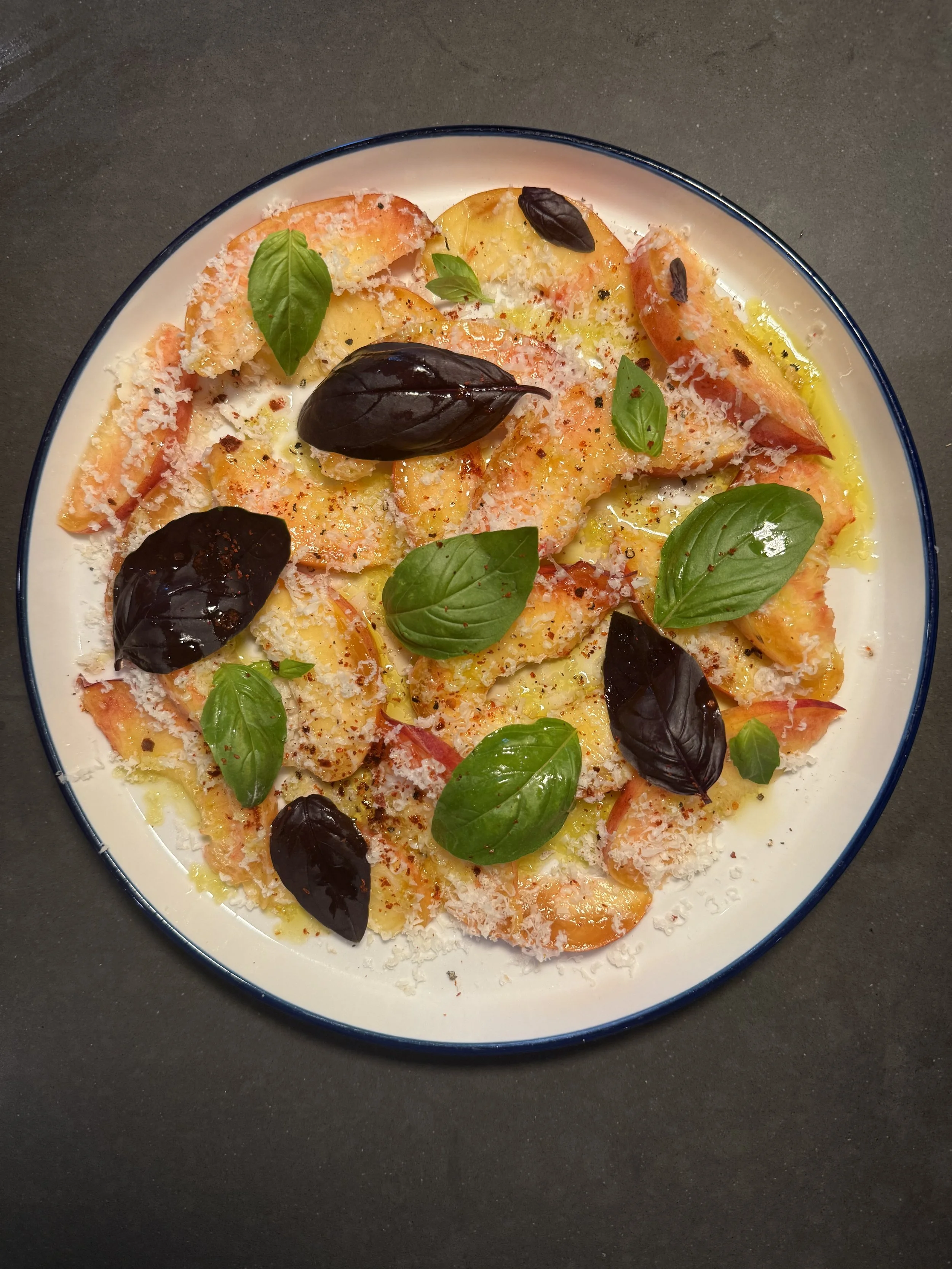 A plate of cooked ravioli pasta topped with grated cheese, fresh basil leaves, black basil leaves, and olive oil on a dark surface.
