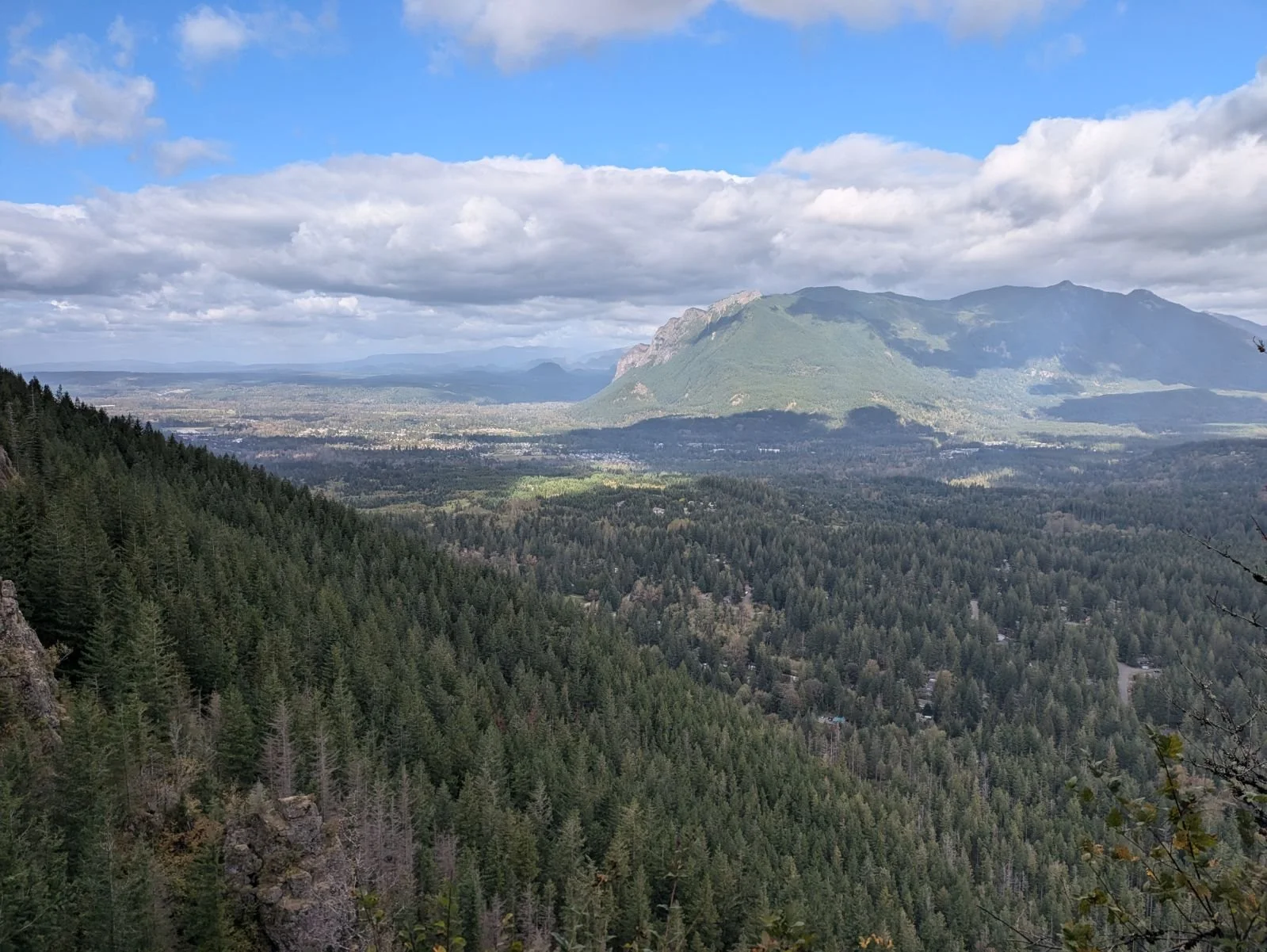 A panoramic view of a forested mountain landscape with a large mountain in the distance under a partly cloudy sky.