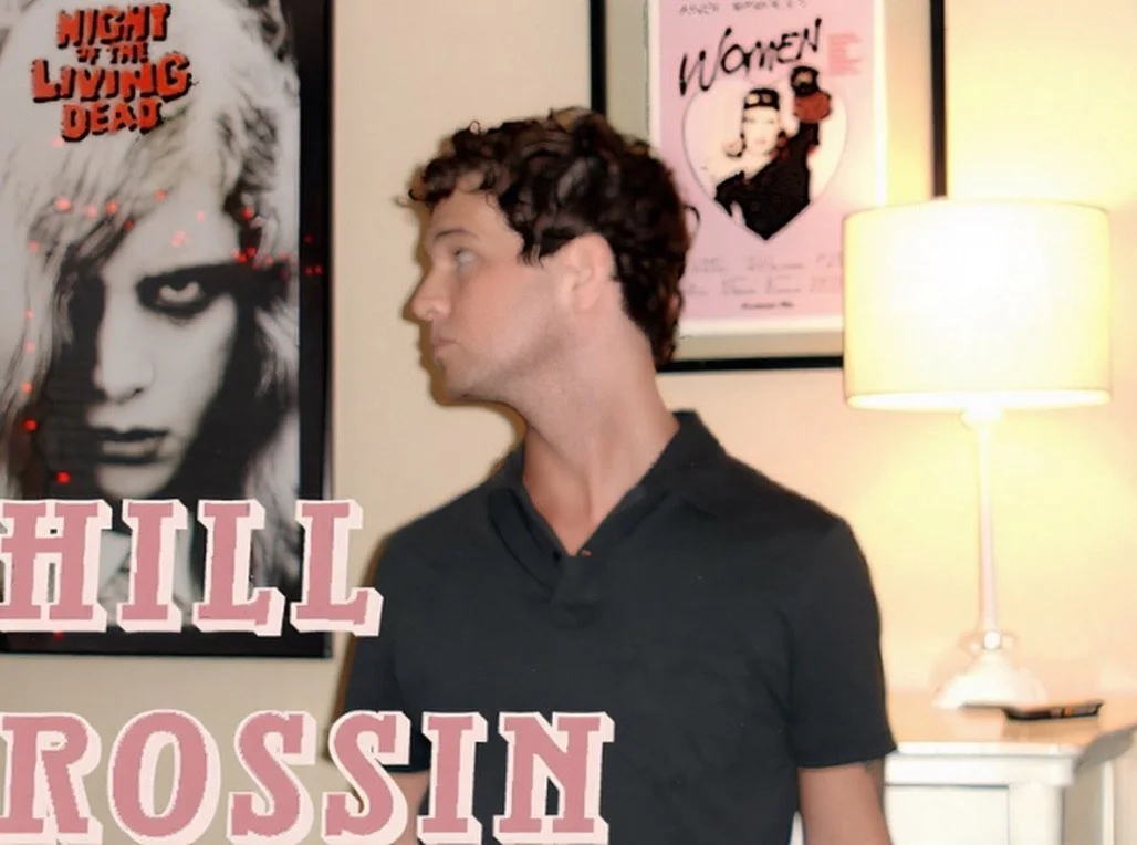 A young man with curly brown hair and a black shirt looking to his left, indoors with posters on the wall and a table lamp nearby.