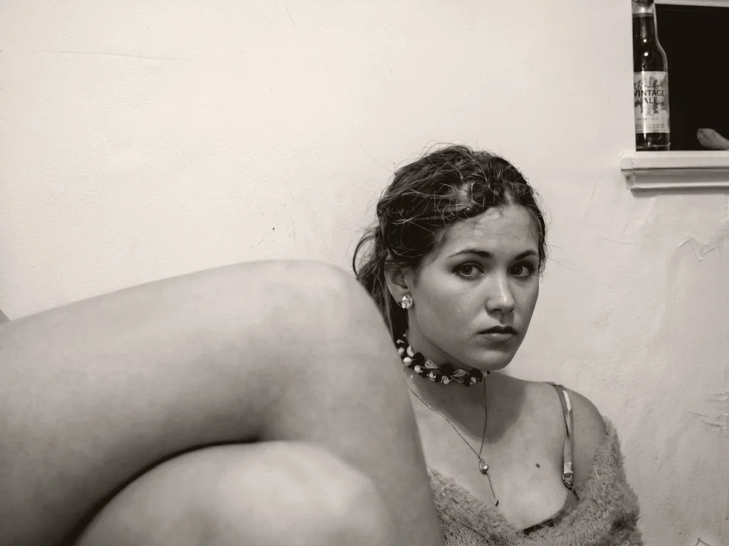 Black and white photo of a woman with short curly hair, wearing earrings, a necklace, and a choker, sitting against a plain wall with a window ledge in the background, partially obscured by a person's shoulder.