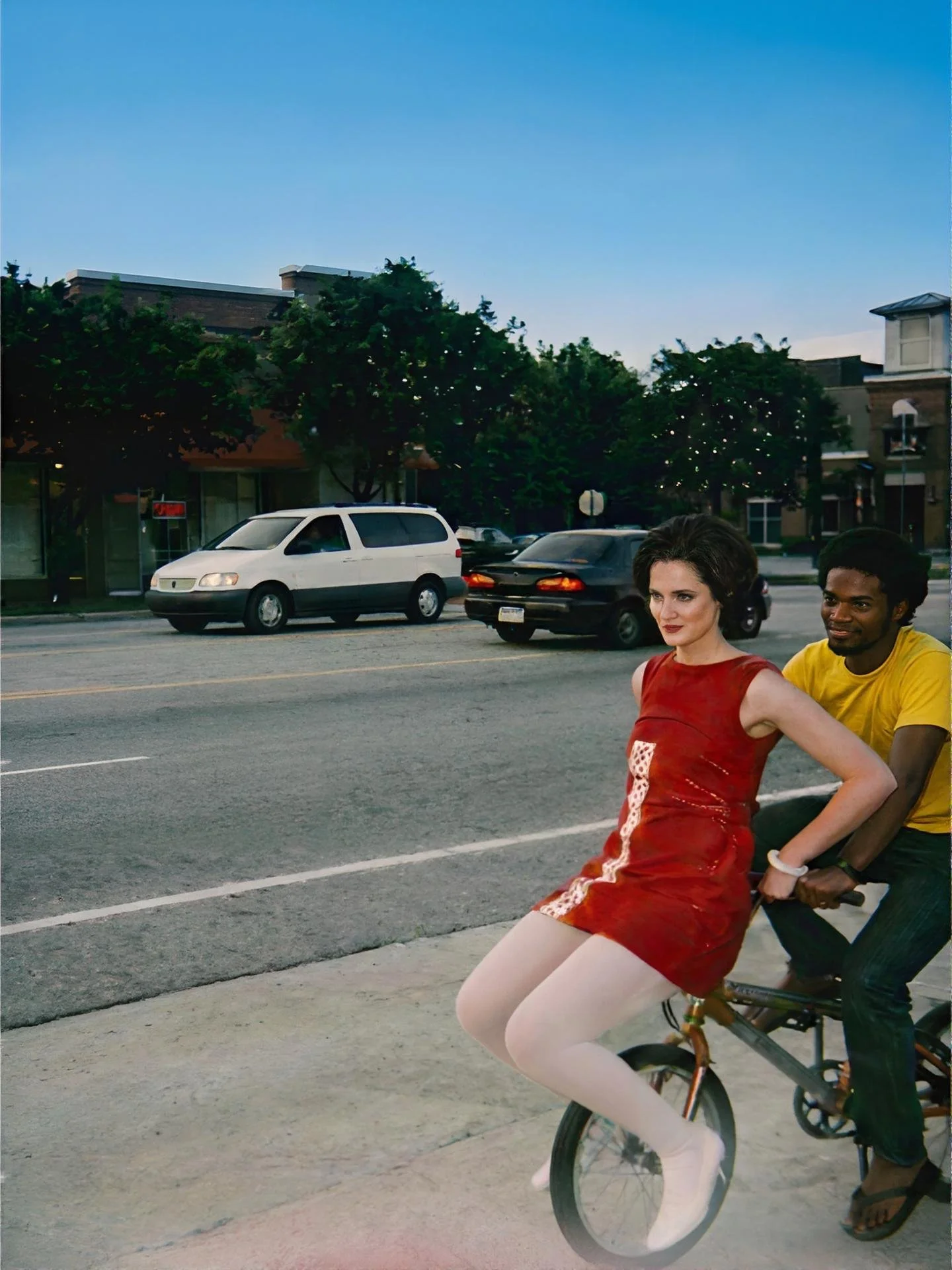 Two people riding a bicycle on a city street during early evening, with parked cars, trees, and buildings in the background.