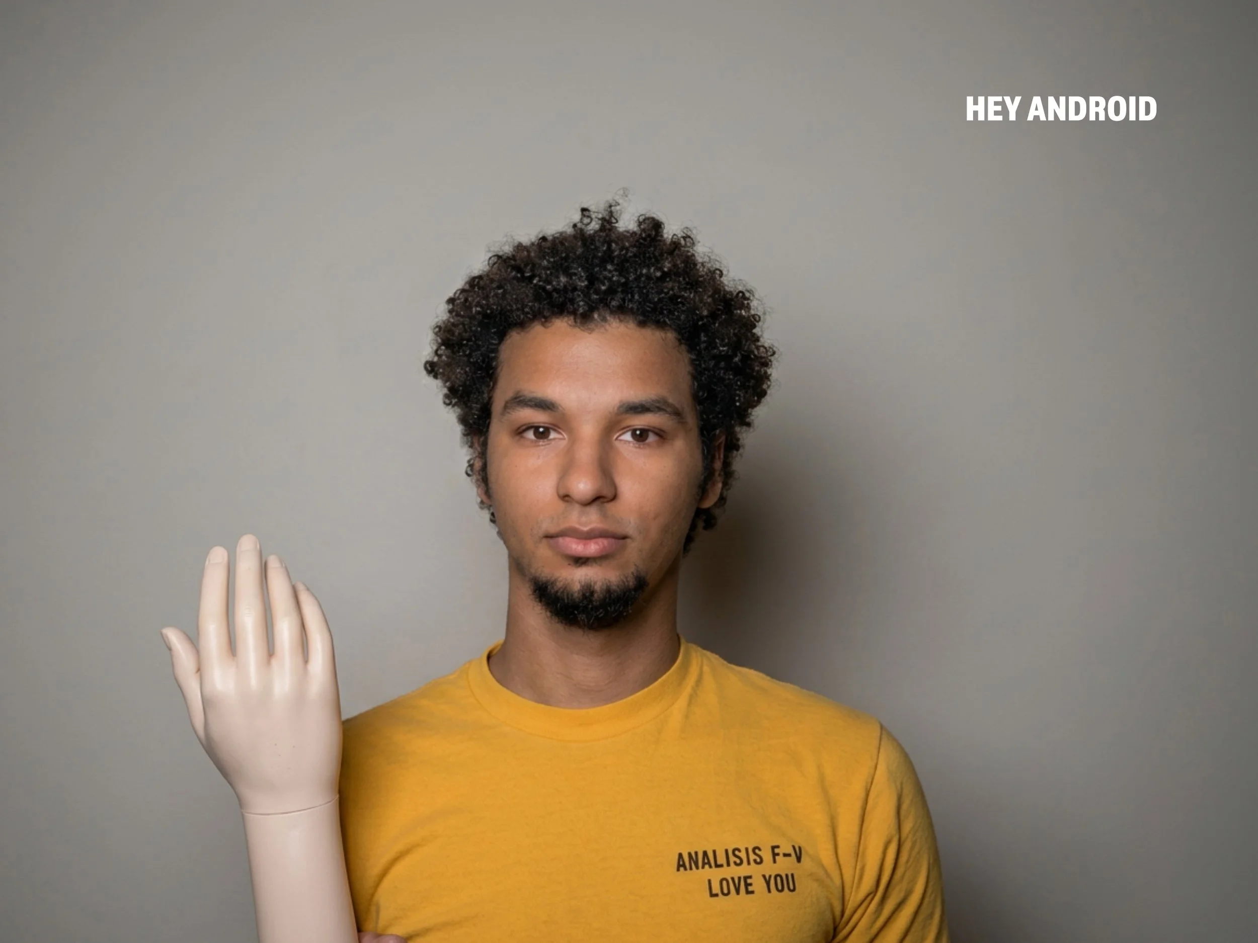 A young man with curly dark hair and a light beard stands against a plain gray background, wearing a yellow T-shirt with black text that reads, 'ANALYSIS F-V LOVE YOU.' He is holding a white mannequin hand up next to his face with the palm facing out