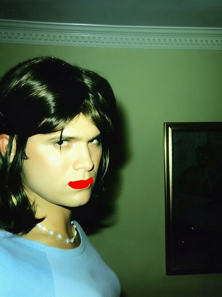 Person with dark hair, wearing a light blue top, red lipstick, and a pearl necklace, facing the camera in a room with green walls and crown molding.