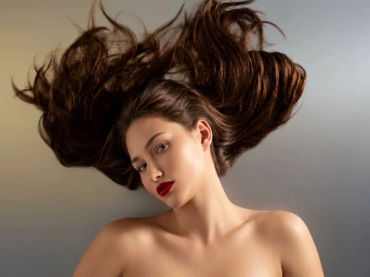A woman with long, flowing brown hair and red lipstick posing with her bare shoulders visible against a neutral background.
