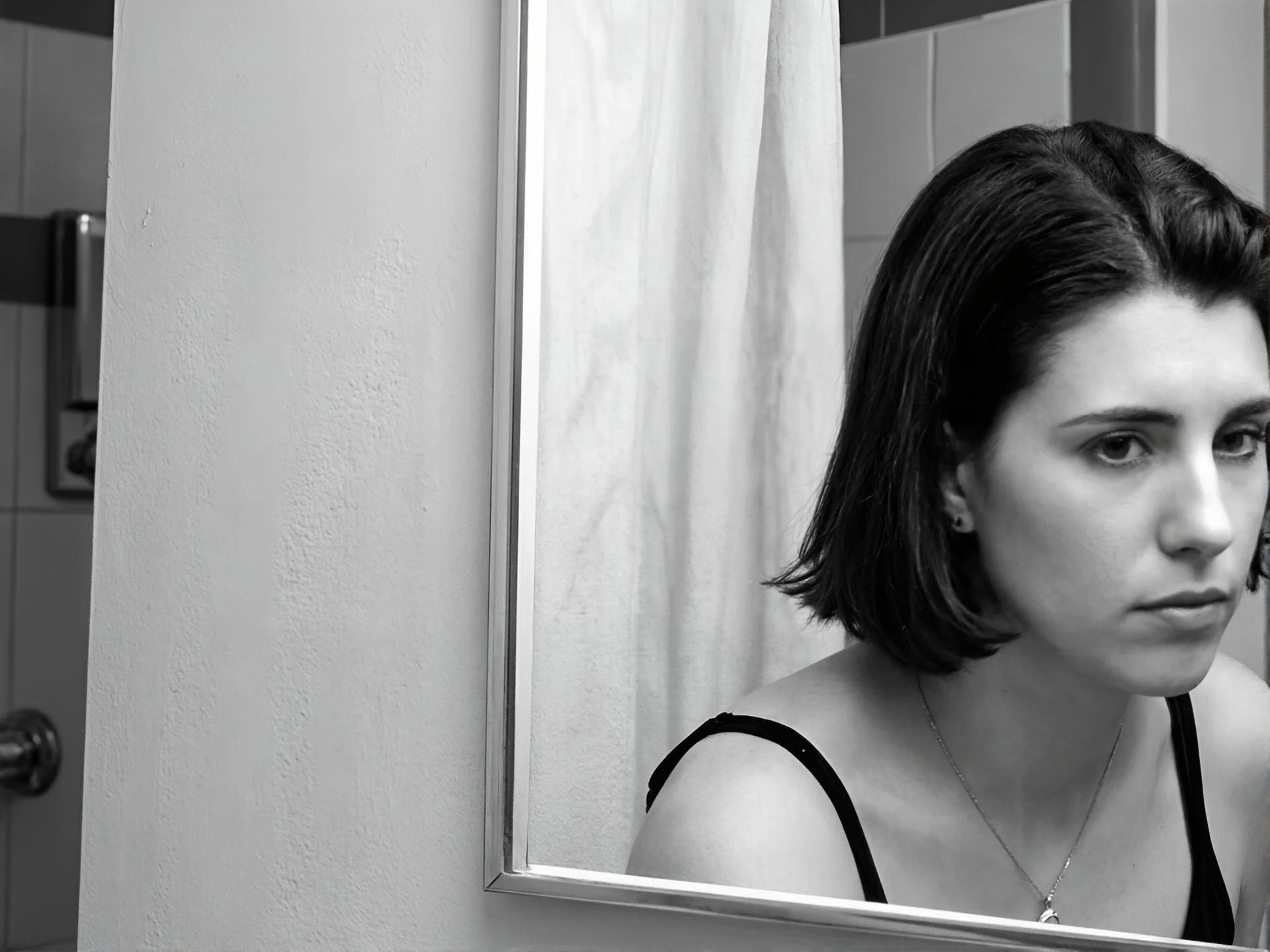 Black and white photo of a woman with short wavy hair looking into a mirror in a bathroom.