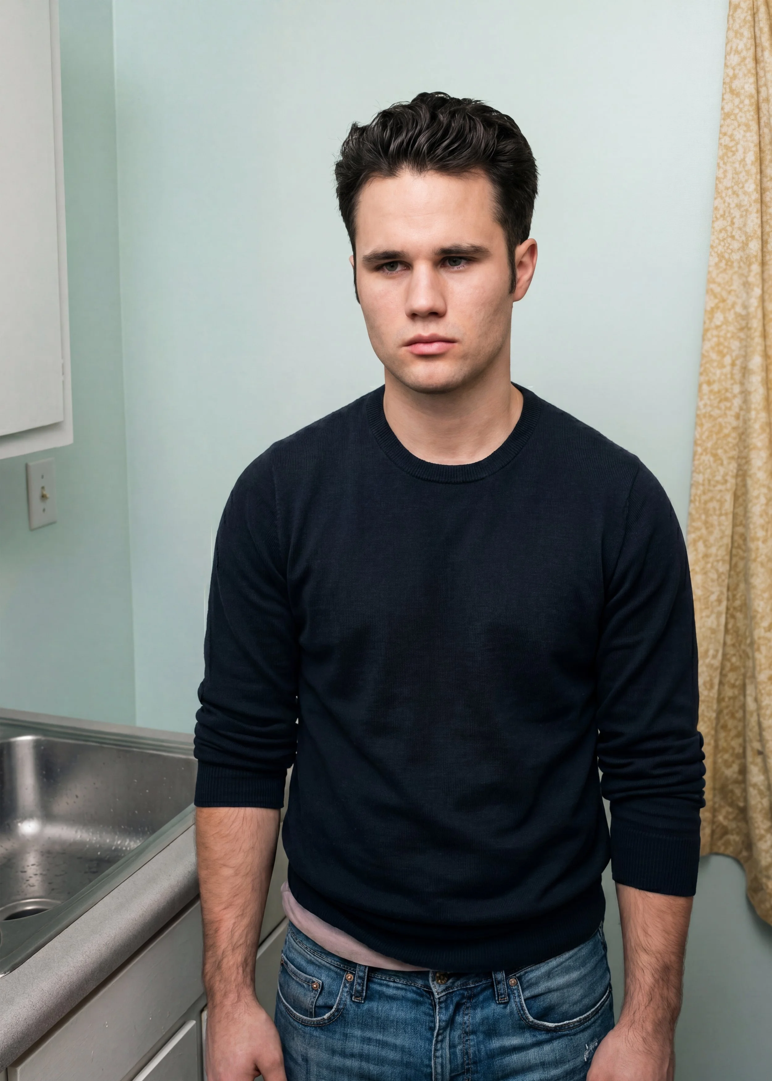 A young man with dark, styled hair stands in a room with light-colored walls and a yellow curtain, wearing a black long-sleeve shirt and jeans, with a serious expression.