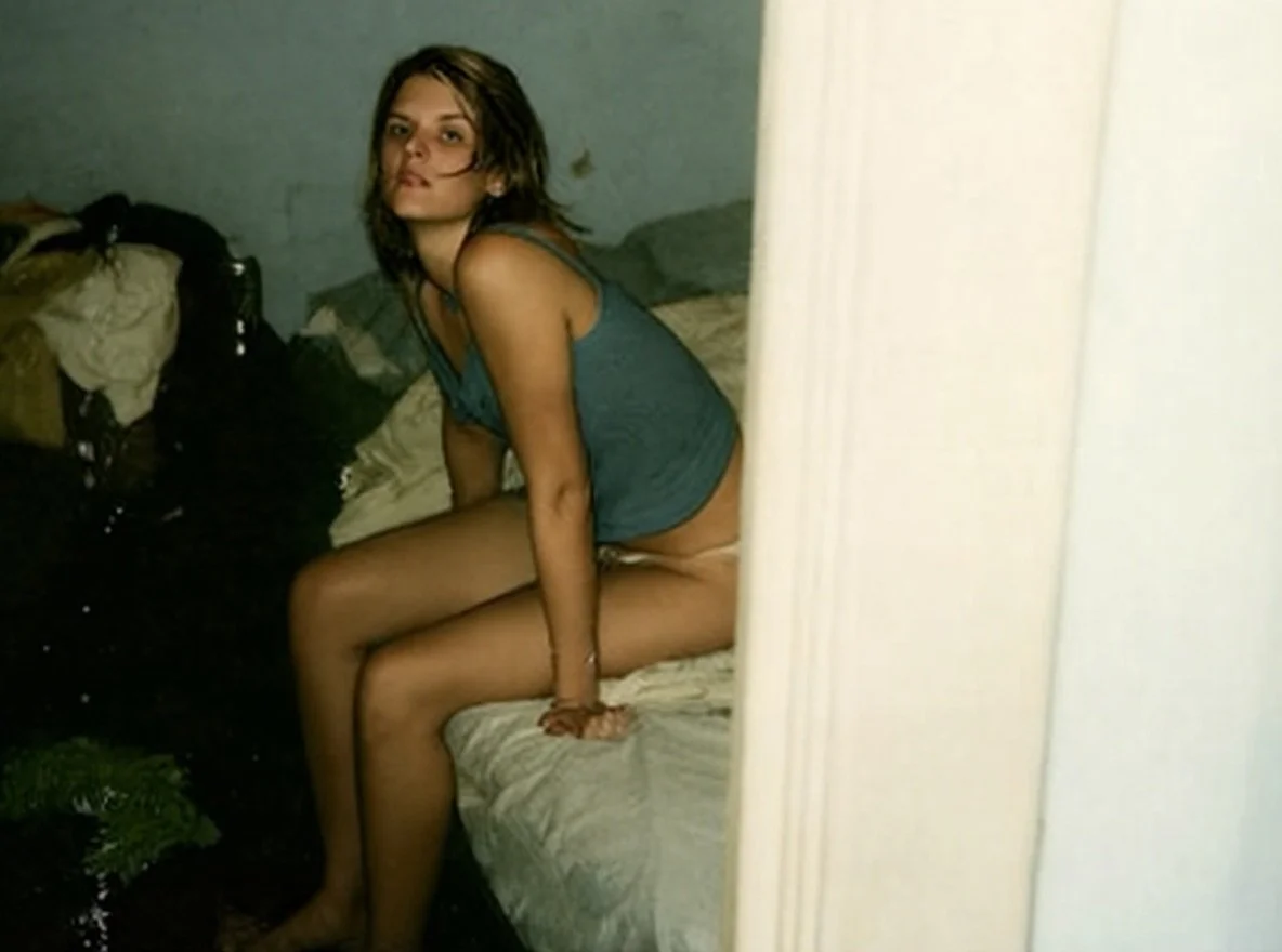 A woman sitting on a bed in a dark room, looking at the camera, wearing a blue tank top, with an unmade bed behind her.