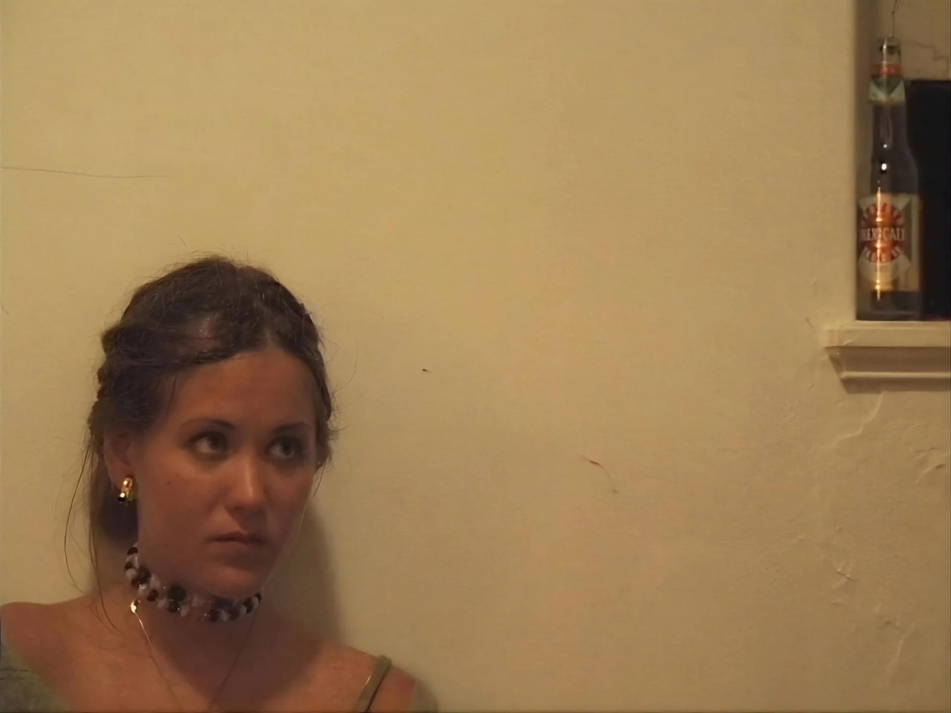 A woman with dark hair tied back, wearing gold hoop earrings, a necklace, and a choker, looks to her left. She is standing against a beige wall with a small shelf holding a glass bottle on her right.