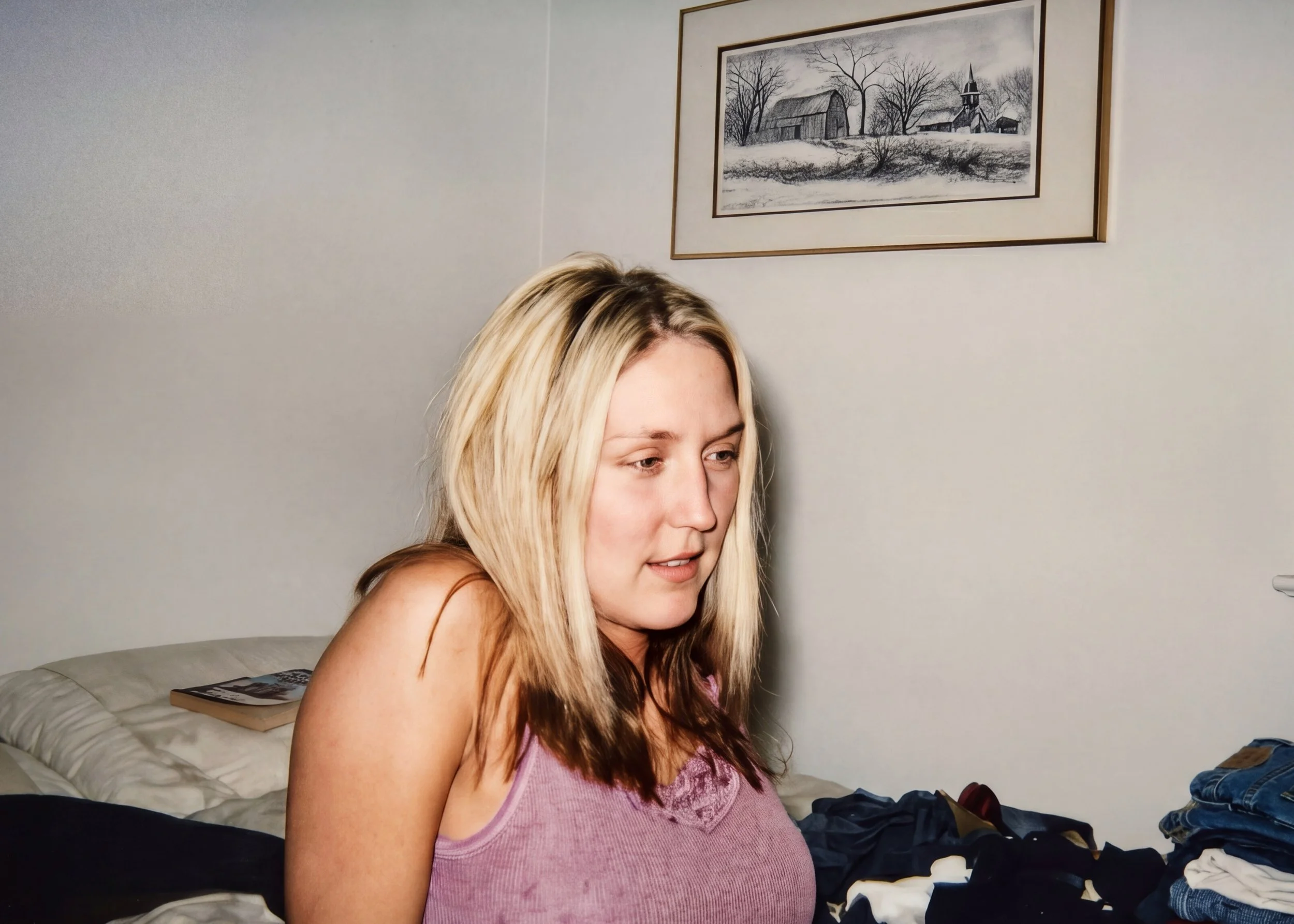 A woman with long blonde hair wearing a pink top is sitting on a bed with clothes scattered around. Behind her, a picture of a snowy landscape with barns and trees is hanging on the wall.