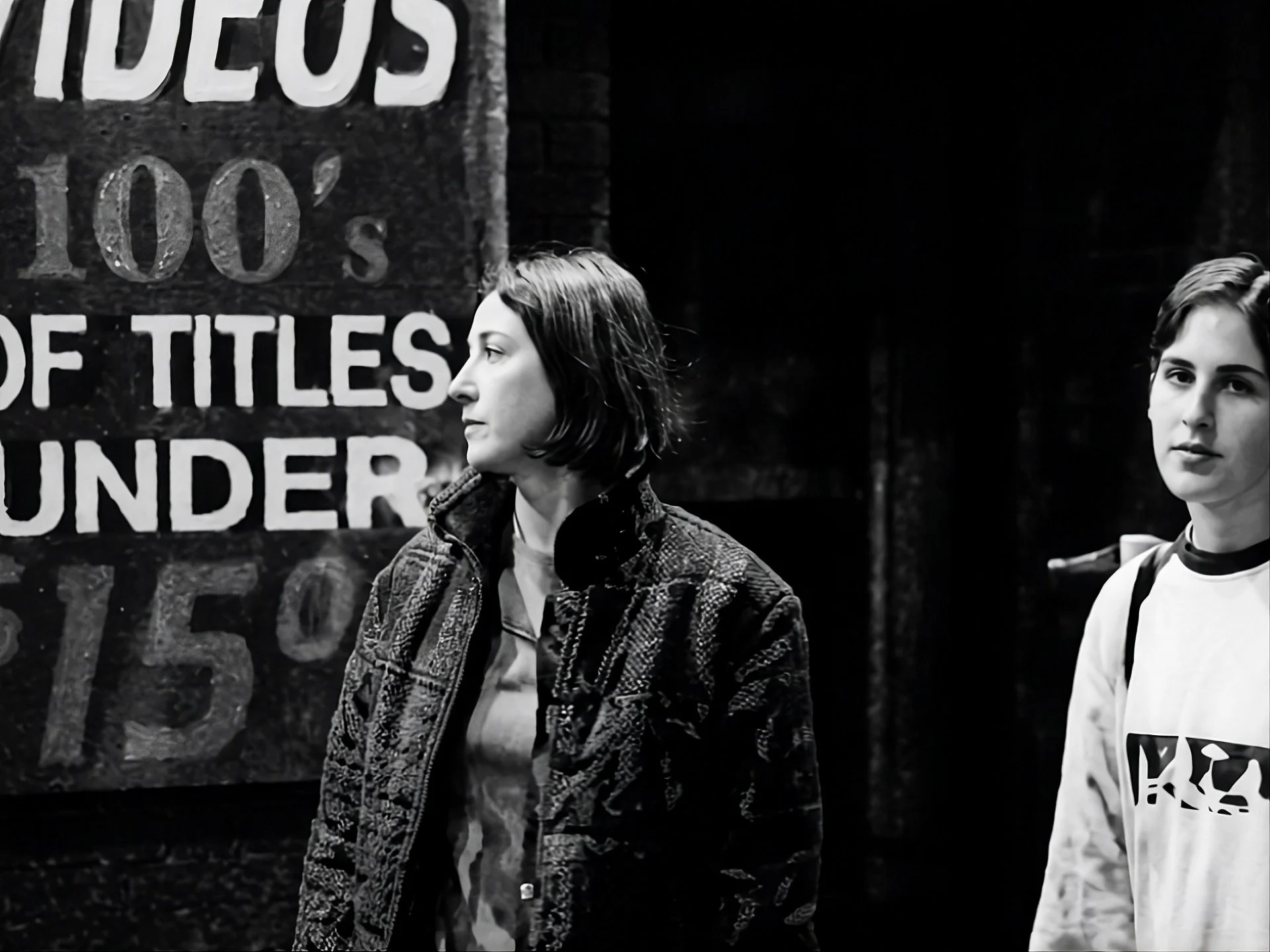 Black and white photo of two young women standing near graffiti-covered wall, one with shoulder-length hair wearing a plaid jacket, and the other with short hair in a hoodie.