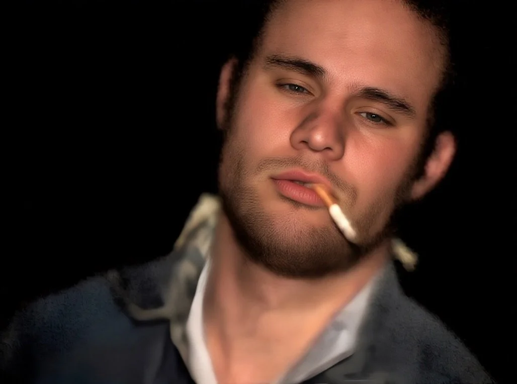 A young man with dark hair and facial hair, wearing a dark jacket, smokes a cigarette, looking into the camera with a relaxed expression against a black background.