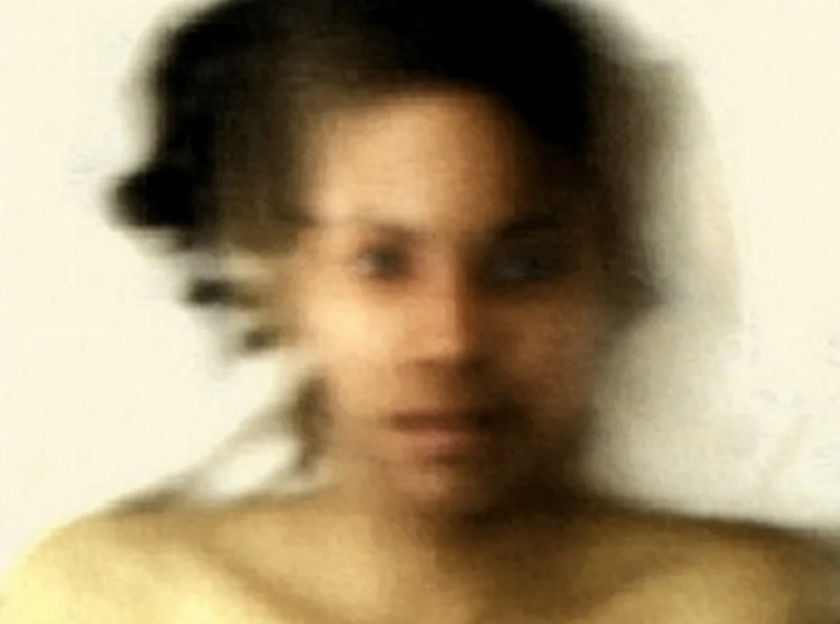 A blurry portrait of a woman with short dark hair and light skin, with a neutral background.