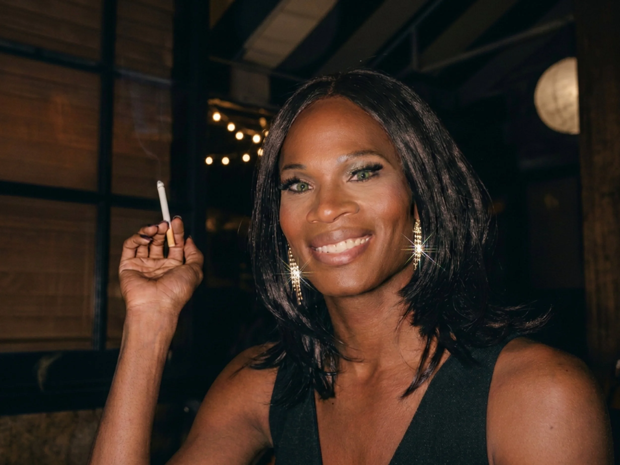 A woman with dark hair and green eyes smiling and holding a cigarette in her hand.