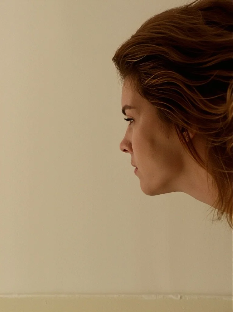 Side profile of a woman with red hair against a plain beige wall.