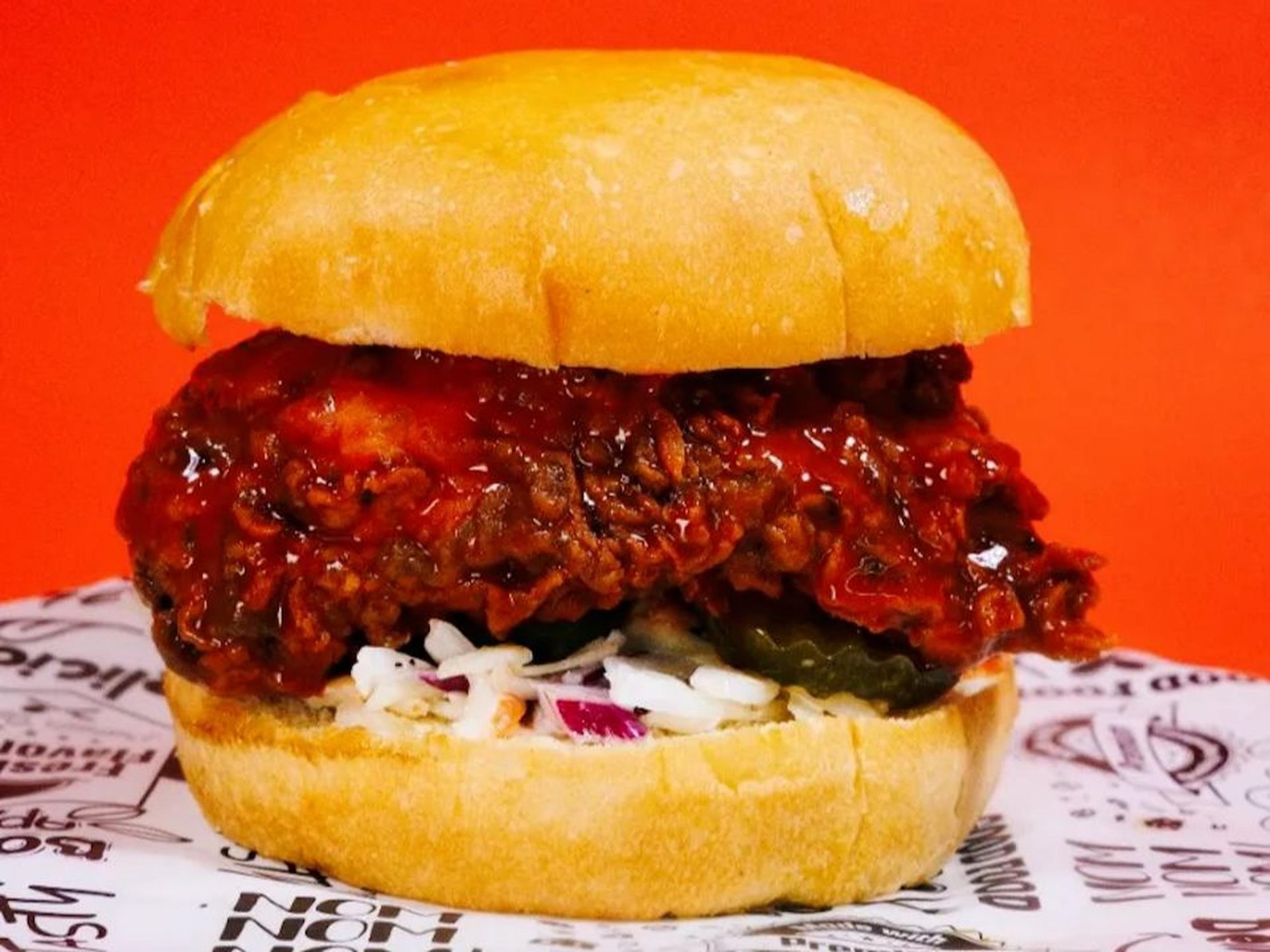 A burger with a toasted bun, shredded lettuce, tomato, pickles, cheddar cheese, onion, a beef patty with sauce, and another bun on top, served on paper with text.