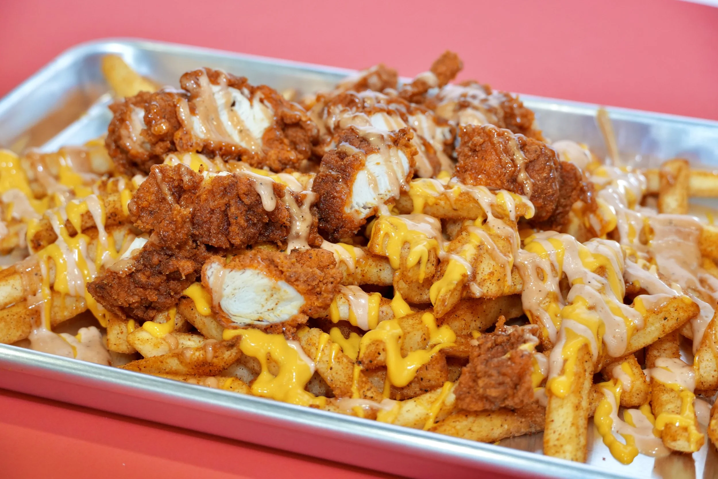 Loaded fries topped with fried chicken pieces, drizzled with cheese and sauce, served in a metal tray on a pink background.