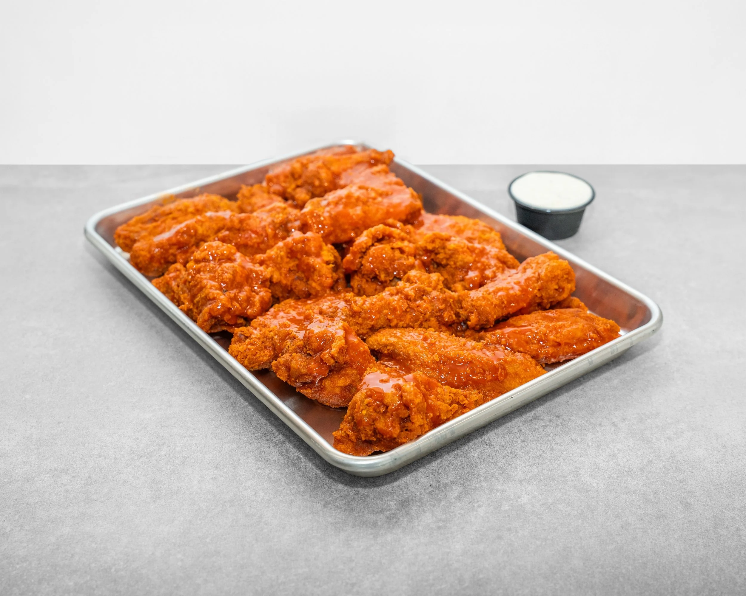 Close-up of a metal tray filled with breaded fried chicken pieces covered in sauce, with a small black cup of creamy dipping sauce on a gray surface.