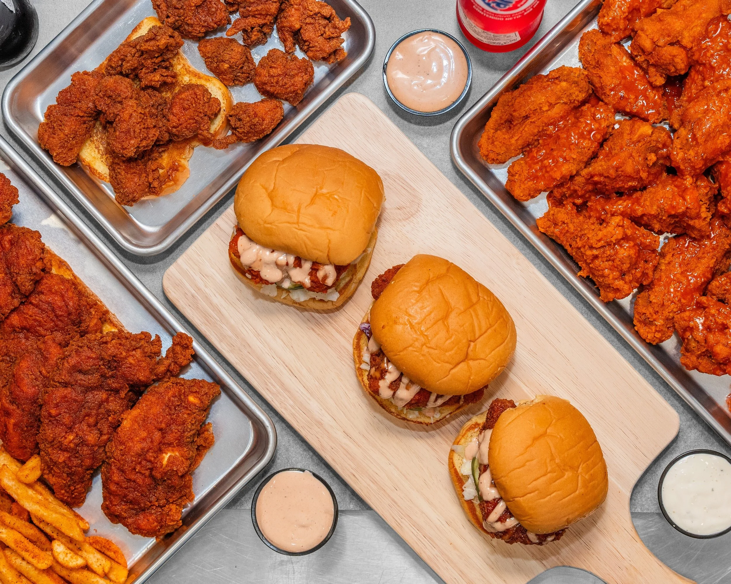 Fried chicken wings, sliders with bun, and French fries on trays, with dipping sauces on a gray table.