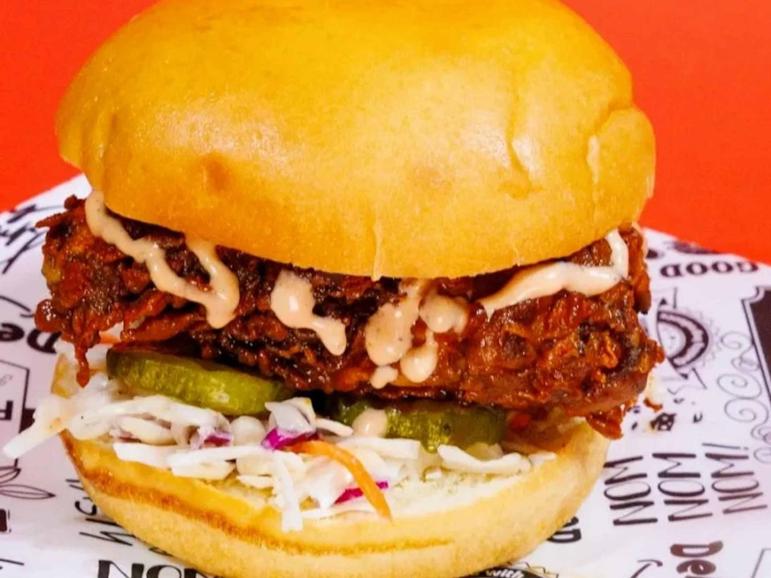Close-up of a sandwich with beef, melted cheese, pickles, cabbage slaw, and a yellow bun against an orange background.