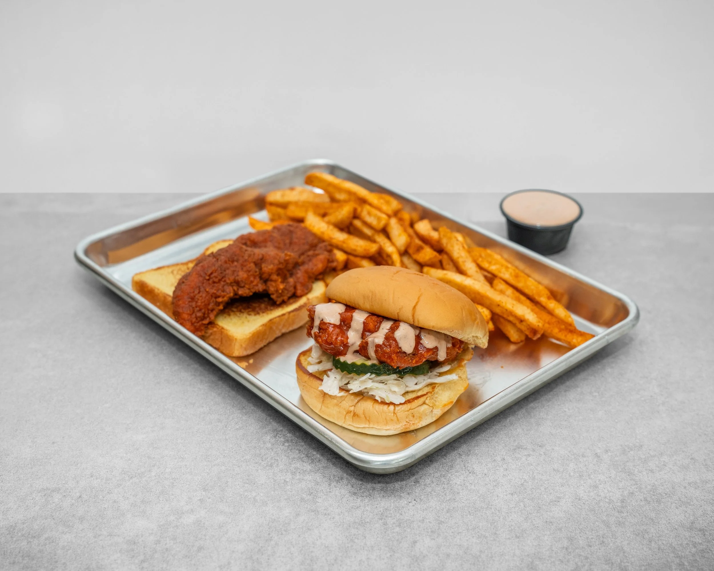Tray of fast food including fried chicken, seasoned fries, a sandwich with chicken, lettuce, pickles, and sauce, and a small container of dipping sauce on a gray surface.