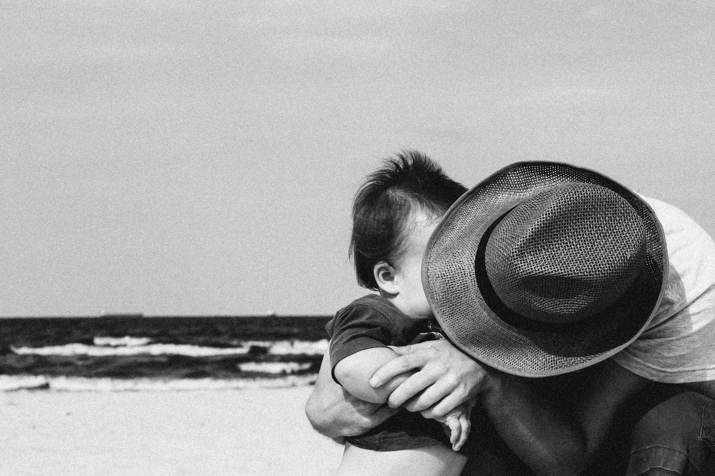 A person sitting on the beach, wearing a large sun hat, with waves in the background, in black and white.