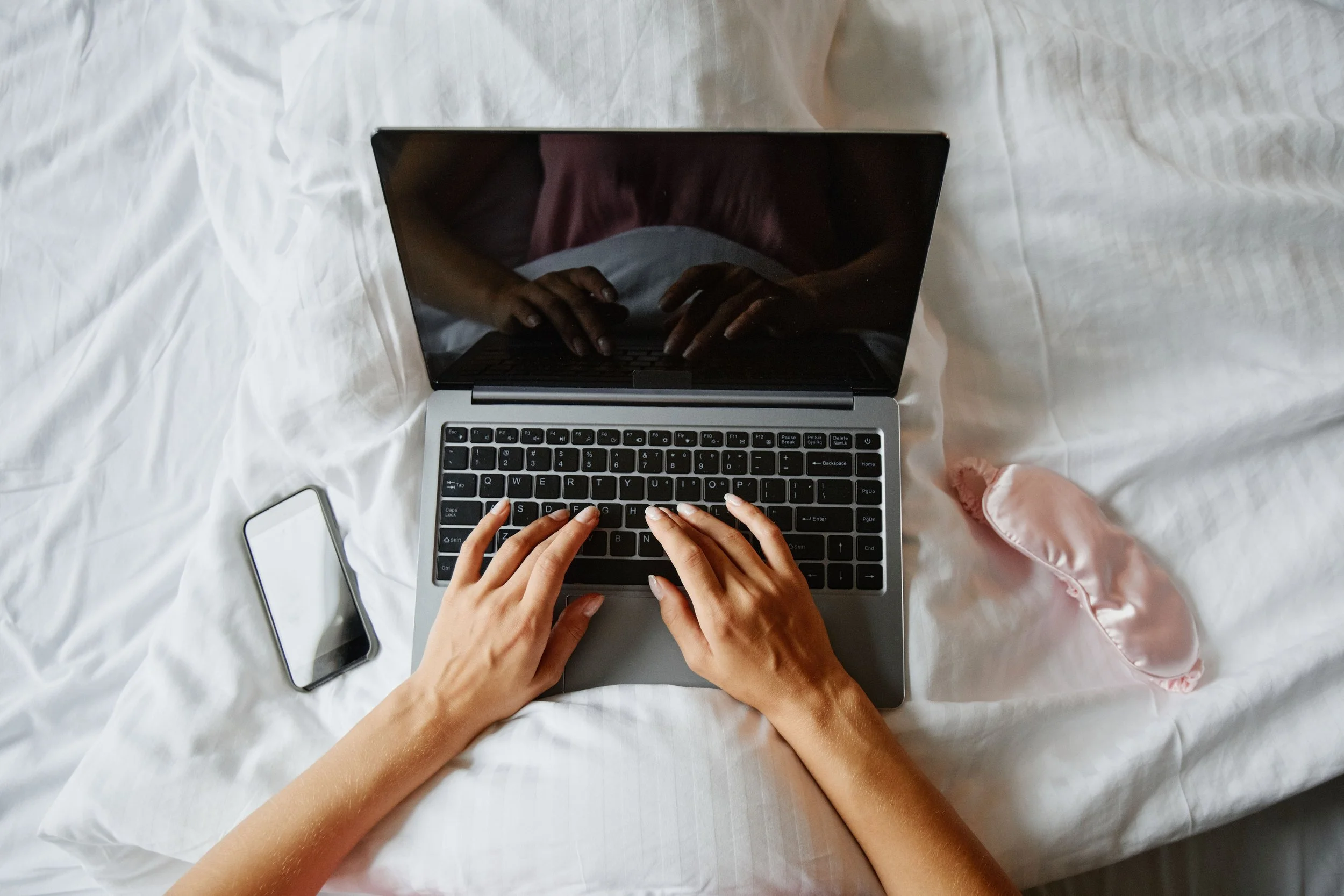 Multiple hands typing on a laptop keyboard on a white bed sheet, with a smartphone and a white charging cable nearby.
