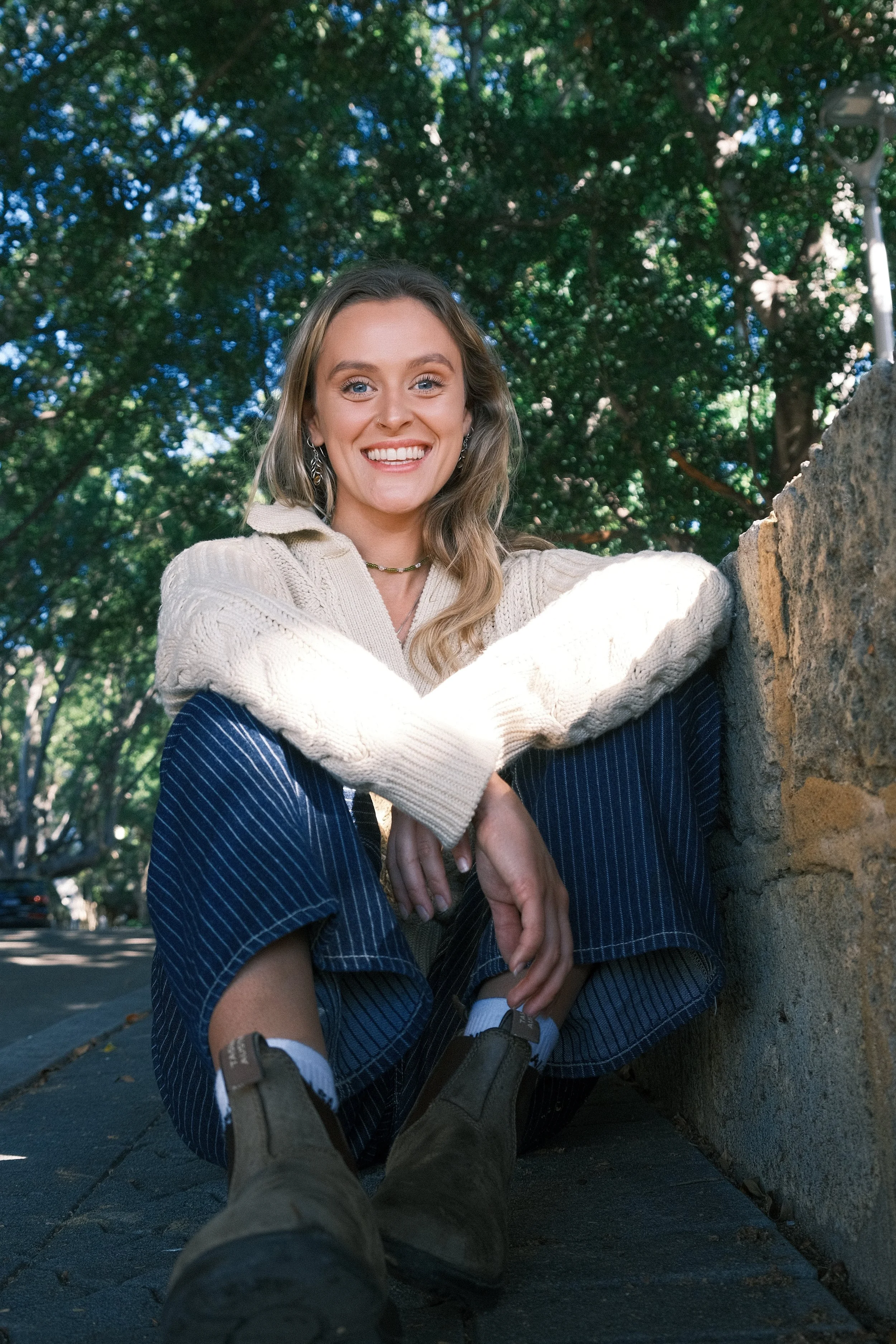 A young woman with blonde hair and blue eyes sitting outdoors near a stone wall, smiling at the camera. She is wearing a cream-colored knit sweater and patterned pants, with trees and blue sky visible in the background.