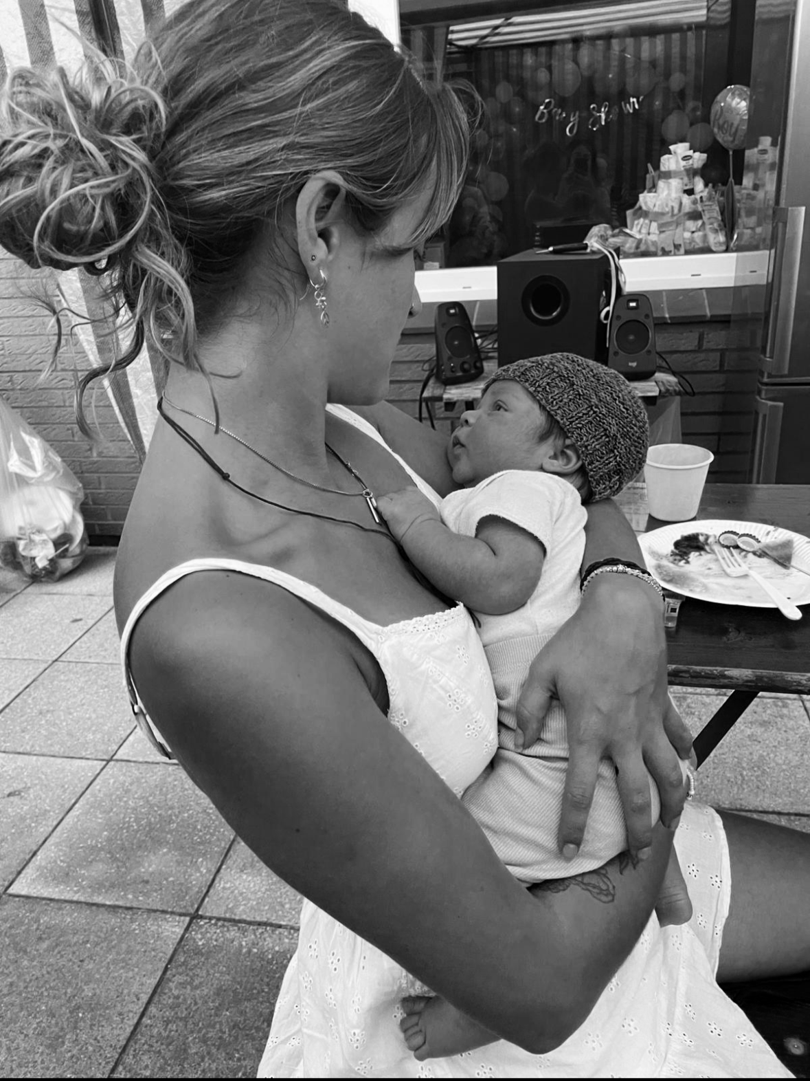 A woman holding a baby in her arms, sitting at a table in a casual indoor setting. The woman has a messy bun hairstyle and is wearing a white dress with eyelet details, earrings, and layered necklaces. The baby, wearing a knit cap and a white outfit, looks up at the woman while she gazes back at the baby. There are plates and utensils on the table, and a speaker and some items on a shelf in the background.
