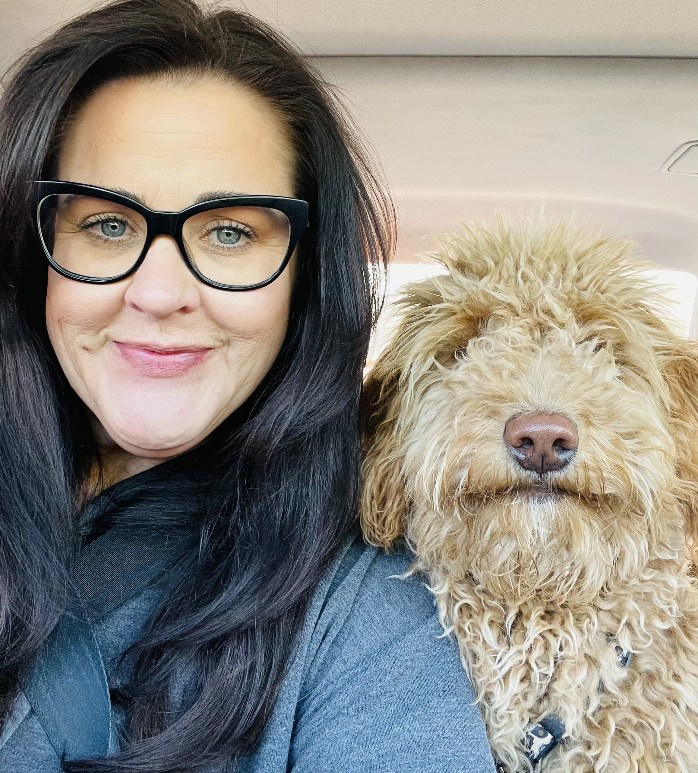 A woman with dark hair and glasses smiling next to a curly-haired dog with light-colored fur. Both are inside a vehicle.
