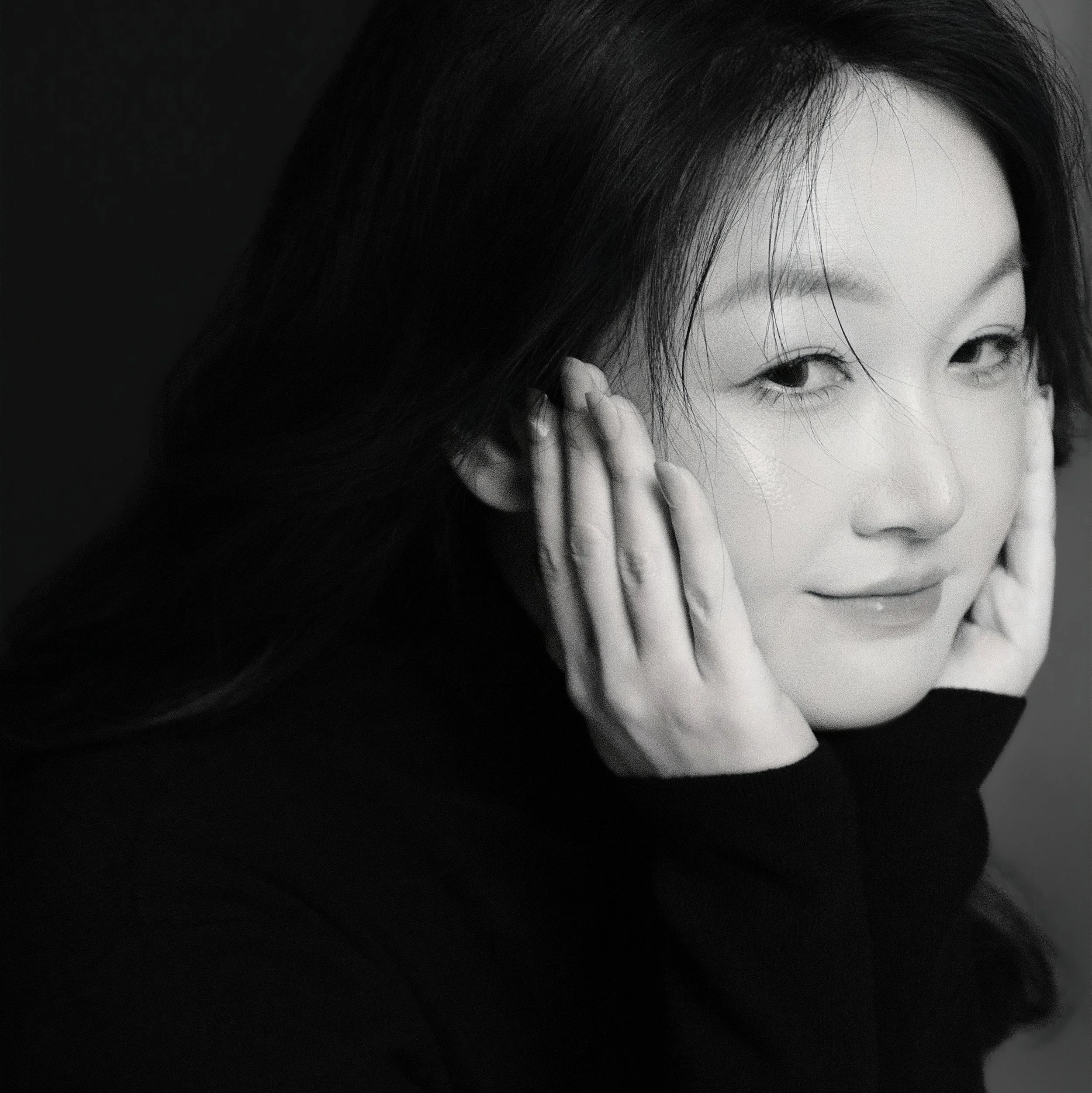 Close-up black and white photograph of a woman's face, focusing on her partially visible eye, eyebrow, and forehead, with her dark hair framing her face.
