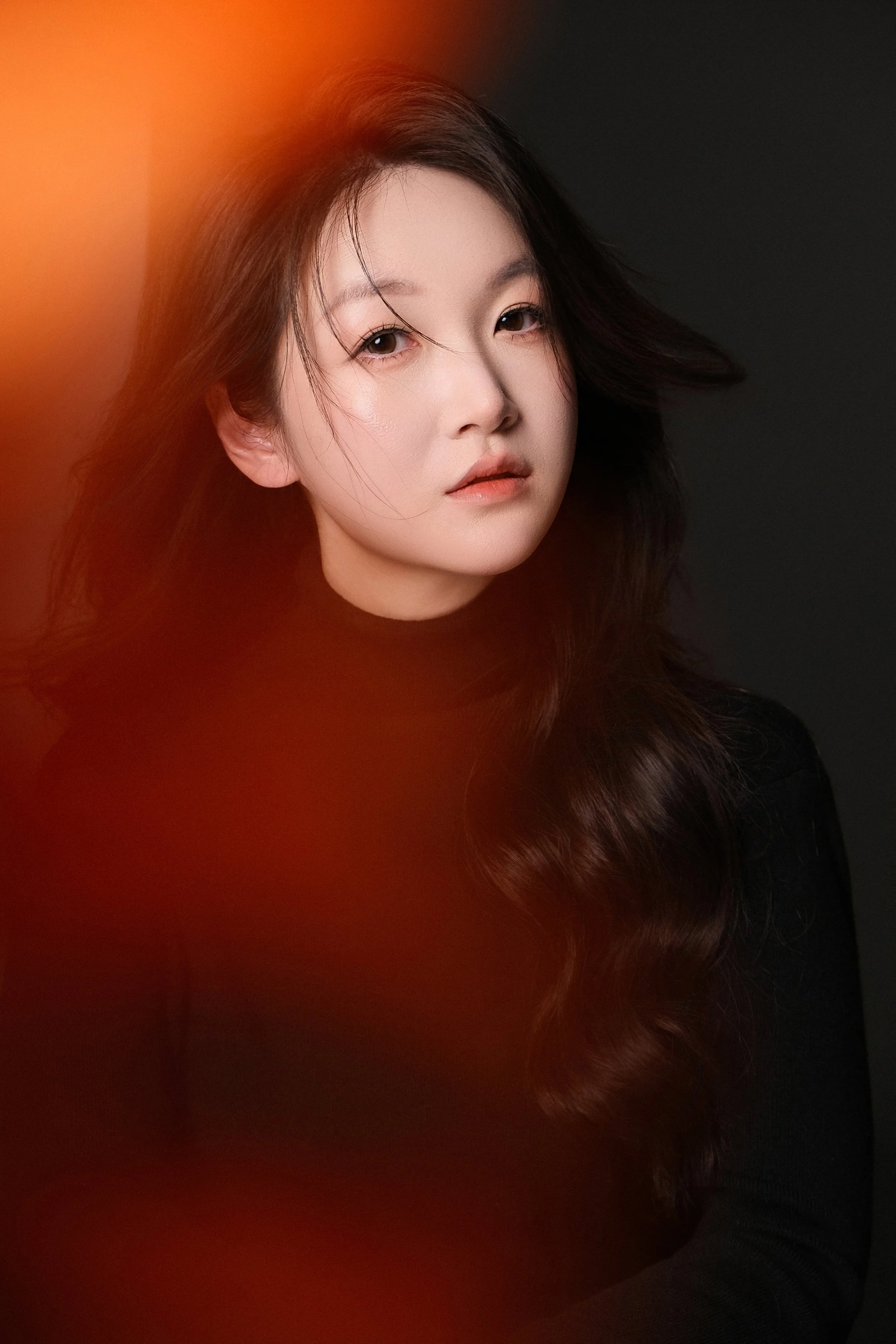 Portrait of a woman with long dark hair, neutral makeup, and wearing a black top, against a dark background with an orange light flare on the left side.