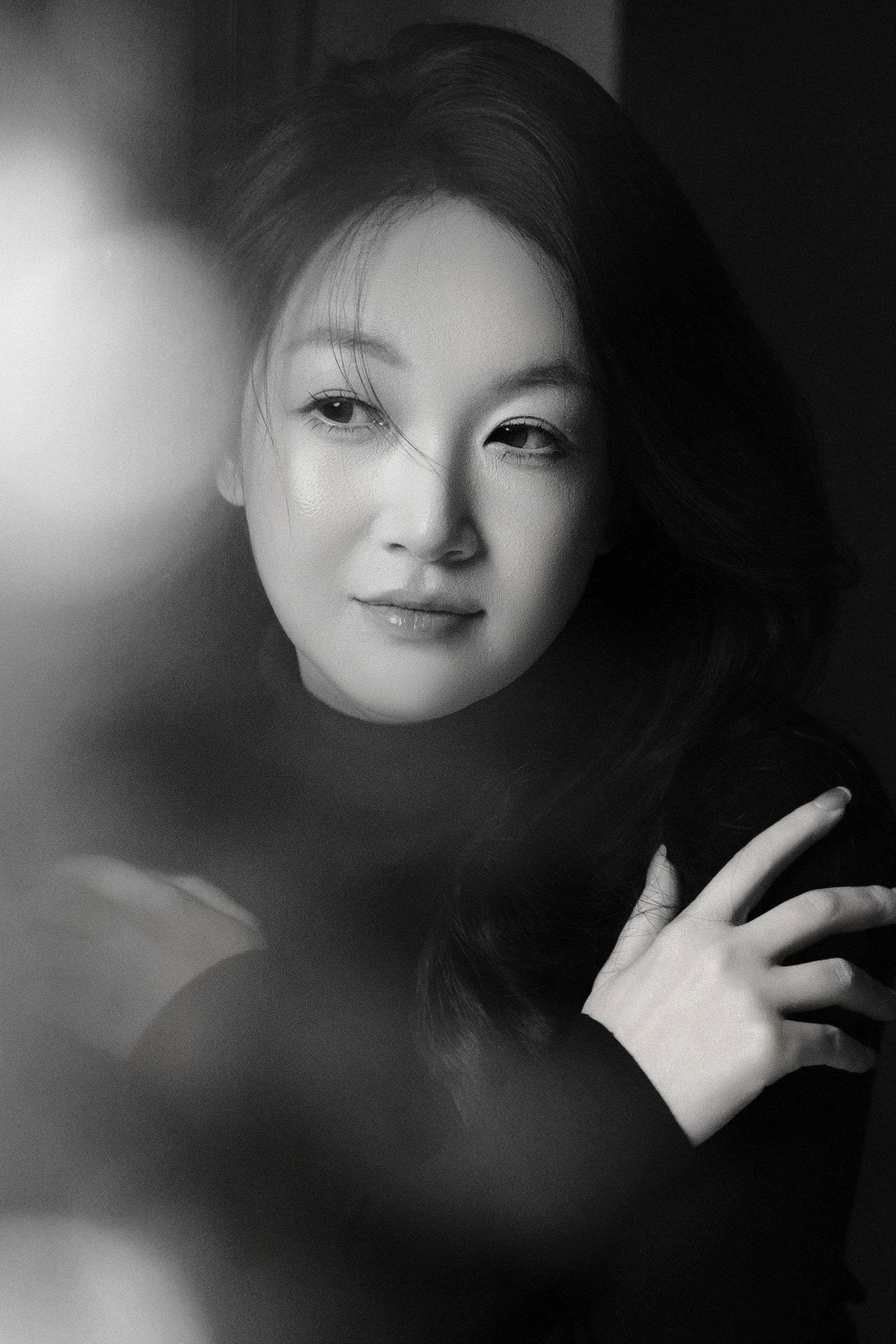 A black and white close-up portrait of a woman with long hair, one eye open and the other partially closed, looking slightly to the side. Her hand is near her face.