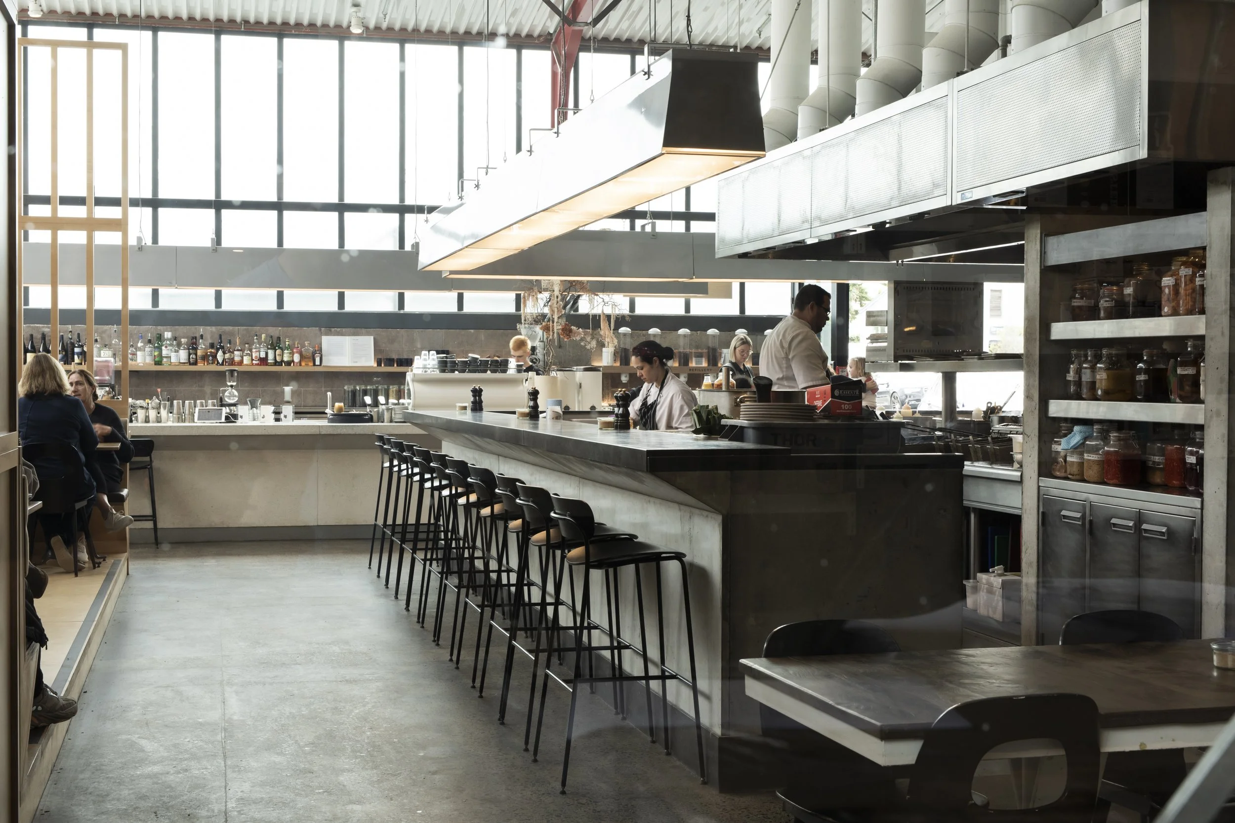 Interior of a modern restaurant or cafe with a high ceiling, large windows, bar seating, and staff working behind the counter.