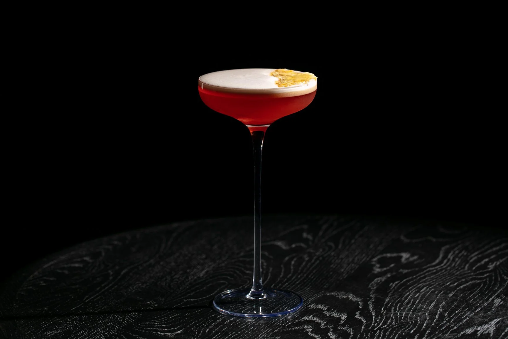 A red cocktail in a coupe glass with a frothy top and a splash of foam or garnish on one side, placed on a dark textured surface against a black background.
