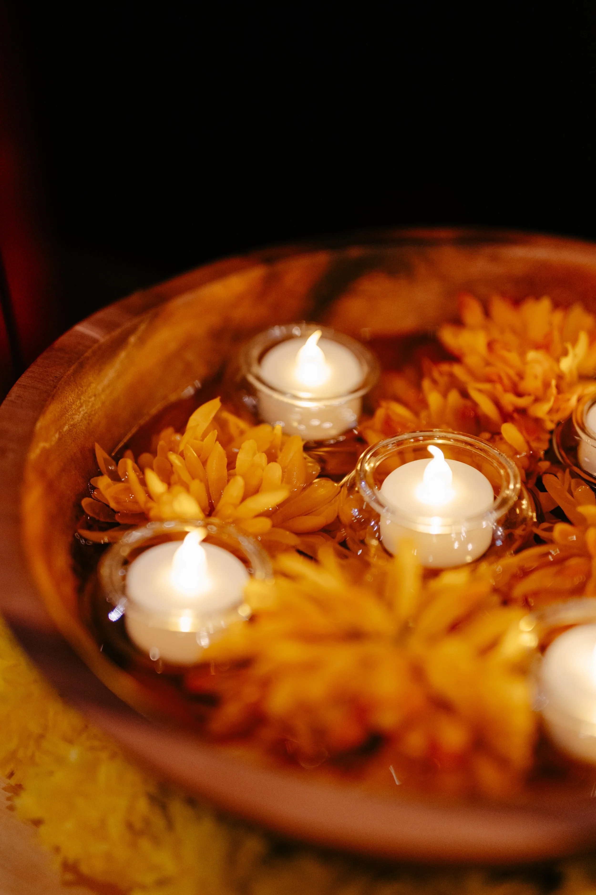 Close-up of a decorative arrangement with yellow flowers and three small candles in glass holders, placed inside a carved wooden bowl.