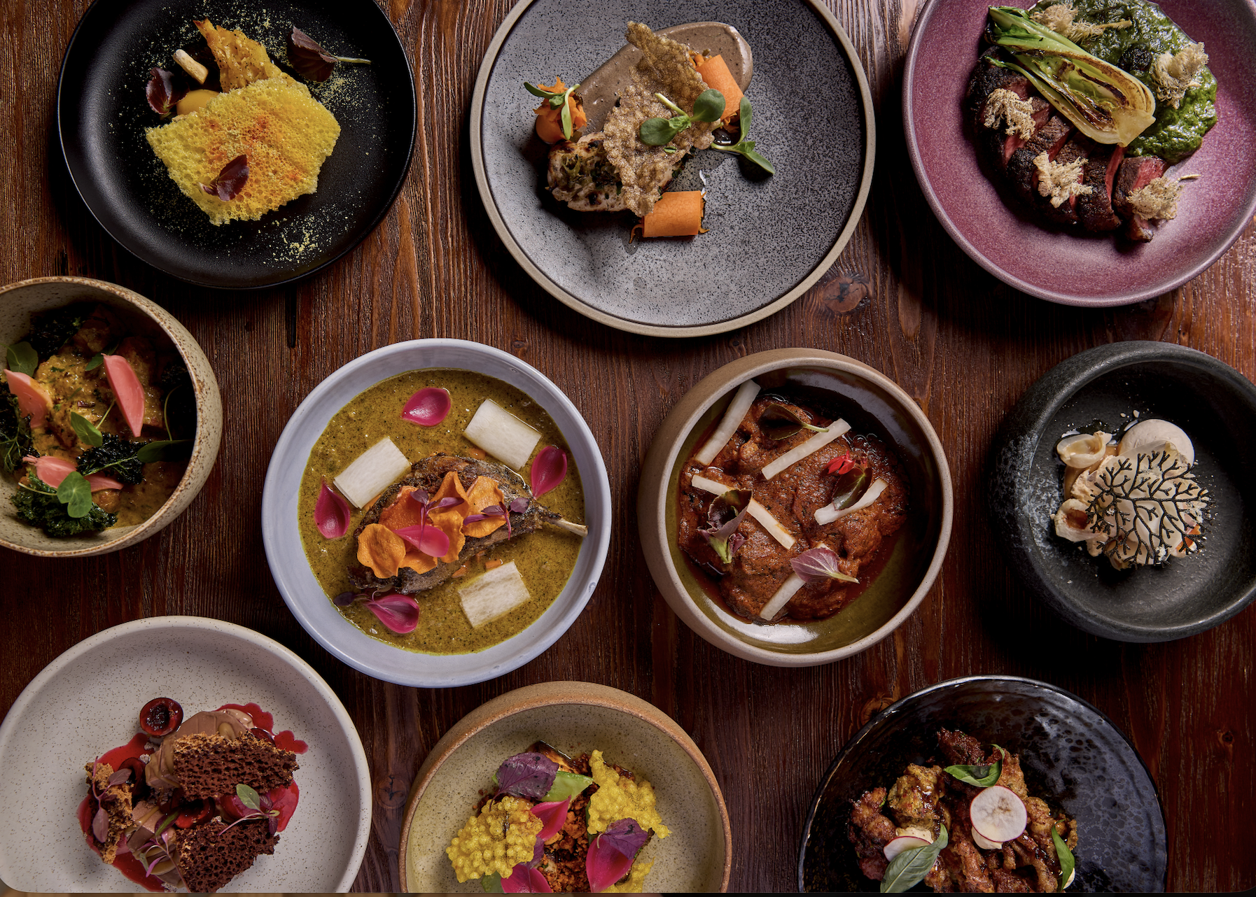 An overhead view of various plated gourmet dishes with colorful garnishes on a wooden table, including meats, vegetables, and sauces.