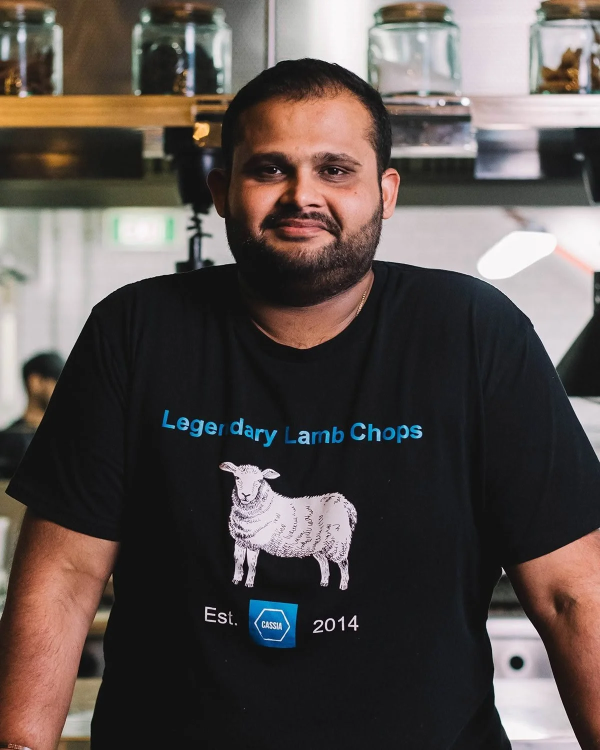 A man with short dark hair, beard, and mustache wearing a black T-shirt with blue and white text and a sheep graphic, inside a restaurant or cafe.