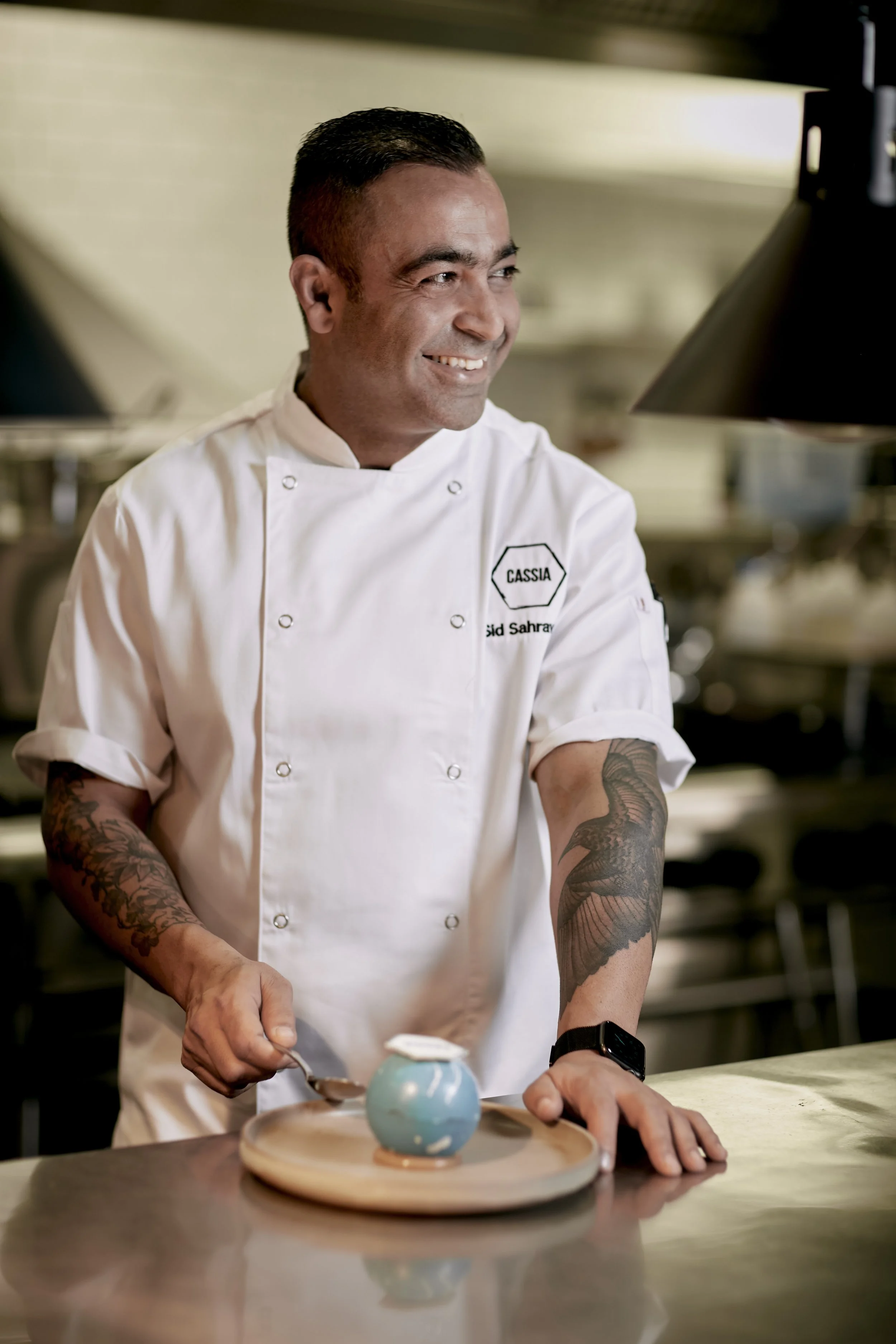 A smiling chef in a white uniform with the name Cassia and the word Saudi Arabia embroidered on it, working in a commercial kitchen. The chef is making a dessert with a blue sphere on a wooden platter.