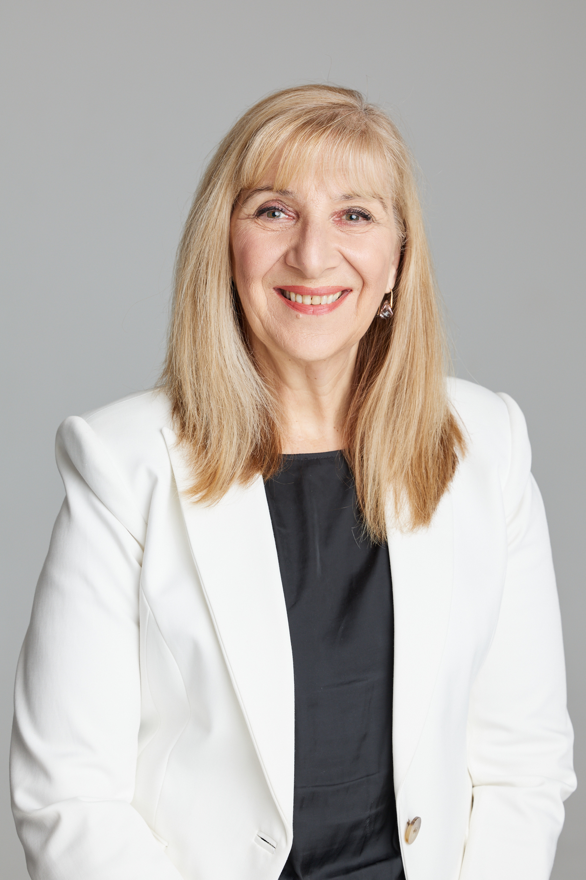 A woman with blonde hair smiling, wearing a white blazer over a black top, against a plain light gray background.