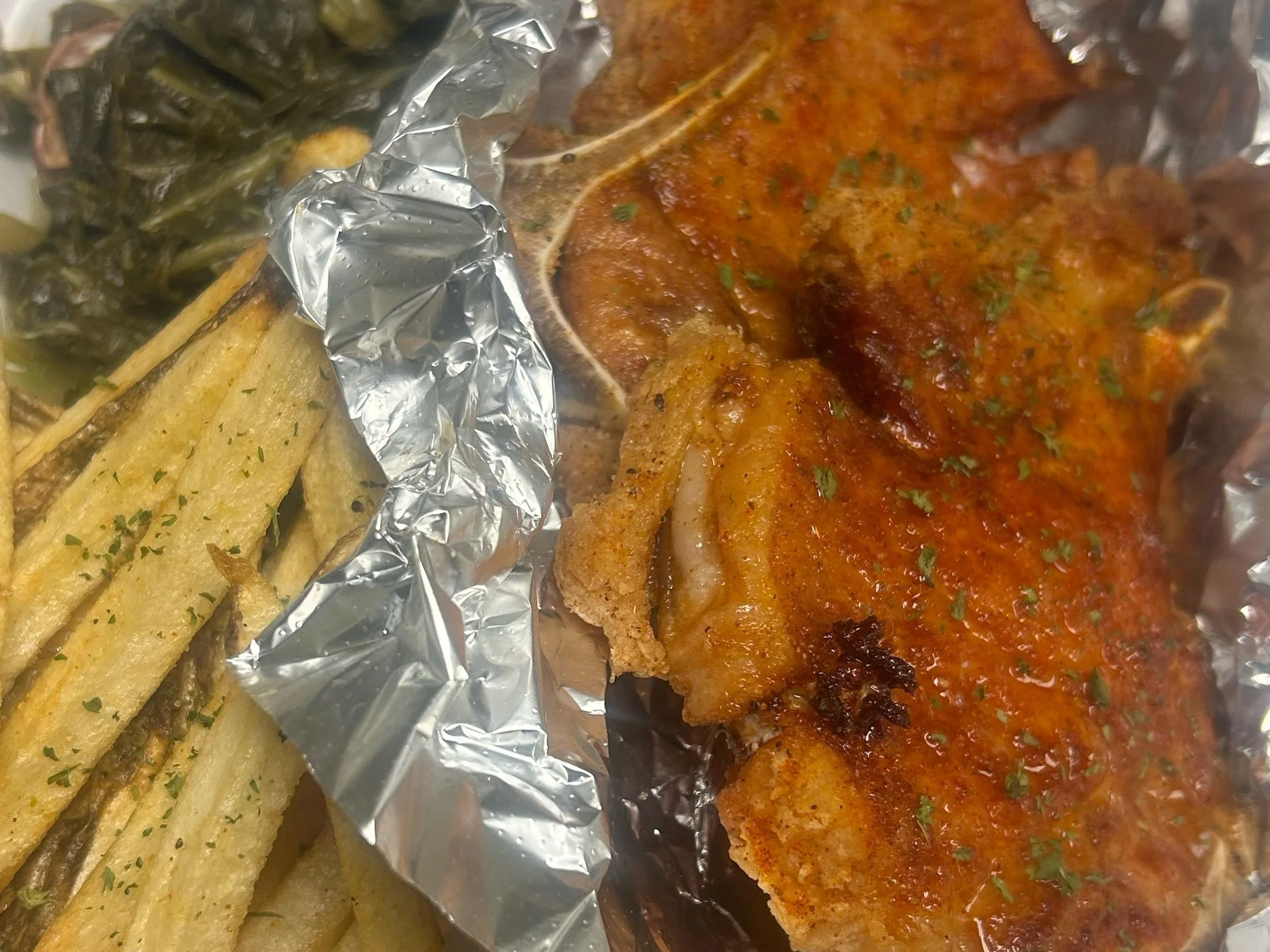 Close-up of baked fish fillet topped with tomato-based sauce and garnished with herbs, alongside crispy seasoned potato wedges on aluminum foil.