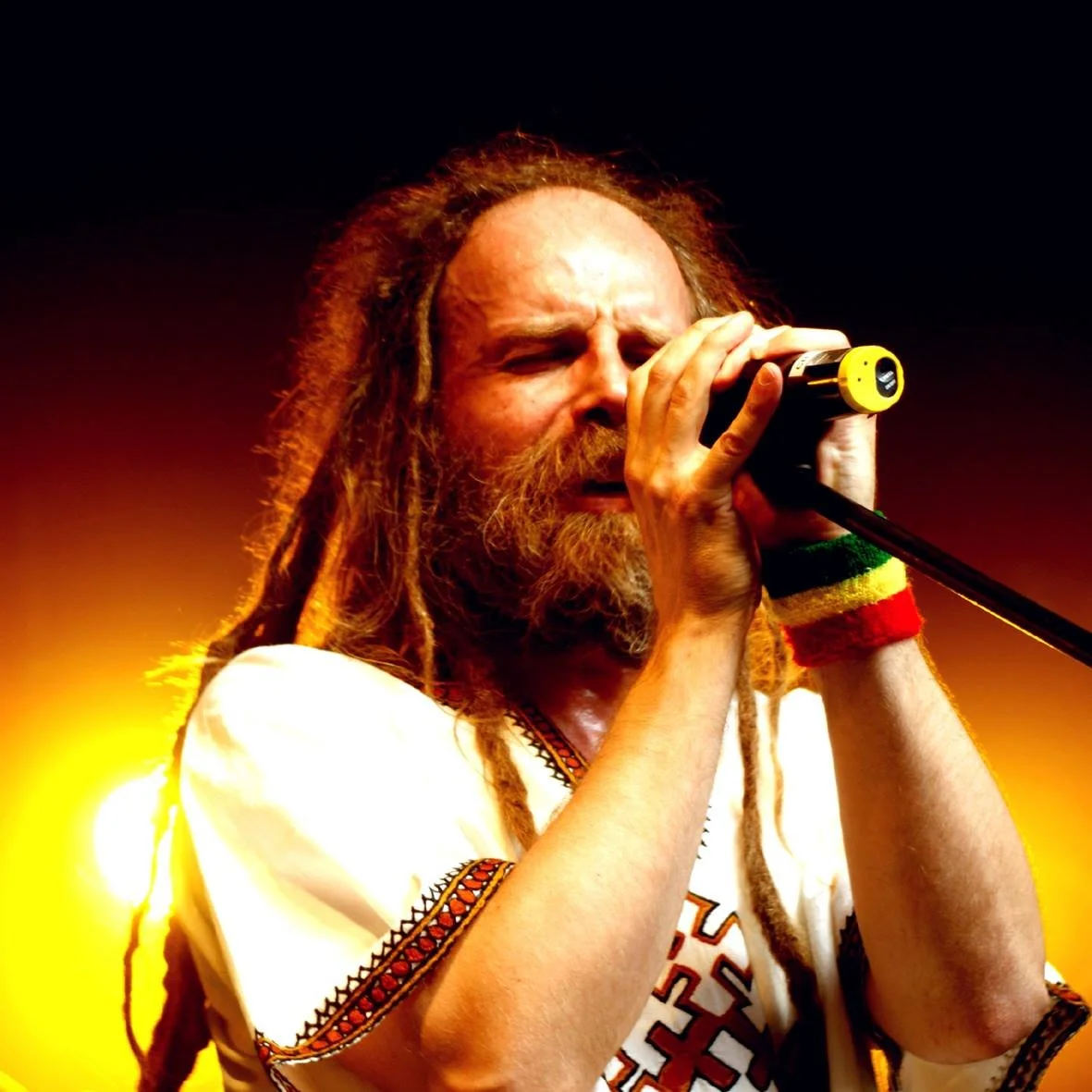 A man with dreadlocks and a beard singing into a microphone, wearing a white shirt with colorful embroidery and a wristband with red, yellow, and green stripes, standing against a dark background illuminated with warm yellow lighting.