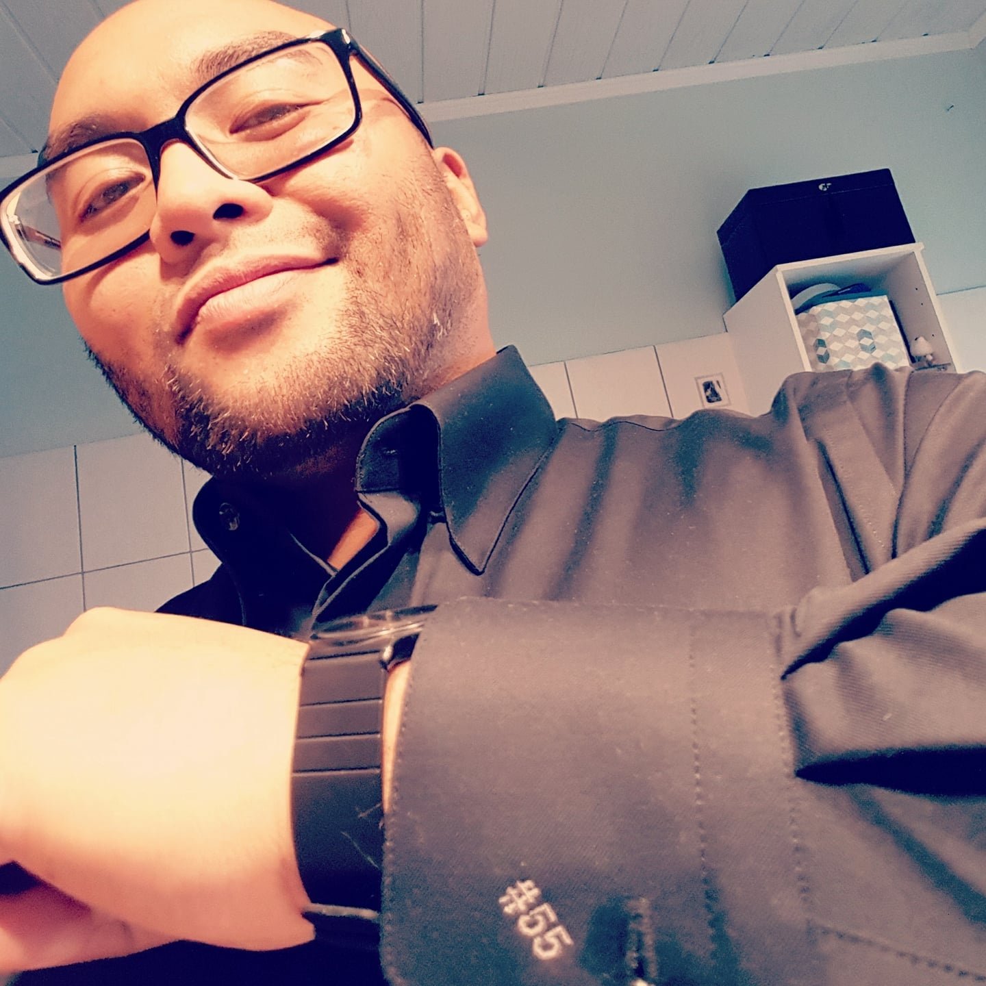 A man wearing glasses, a black shirt, and a watch smiling and taking a selfie in a room with tiled walls and a ceiling with white panels. There is a white shelf with storage boxes in the background.