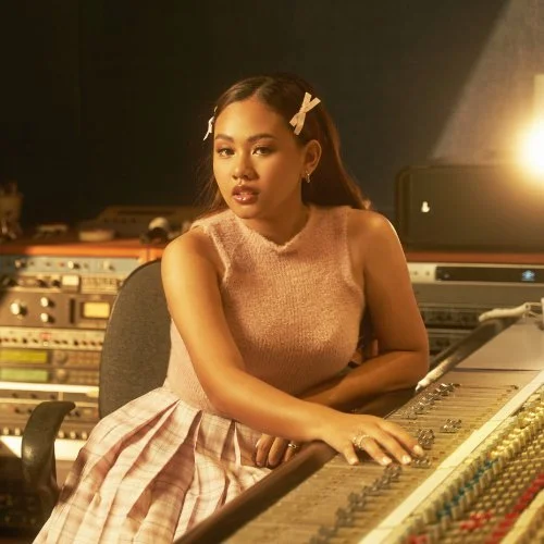 A young woman in a pink dress and hair clips sitting at a mixing console in a recording studio.