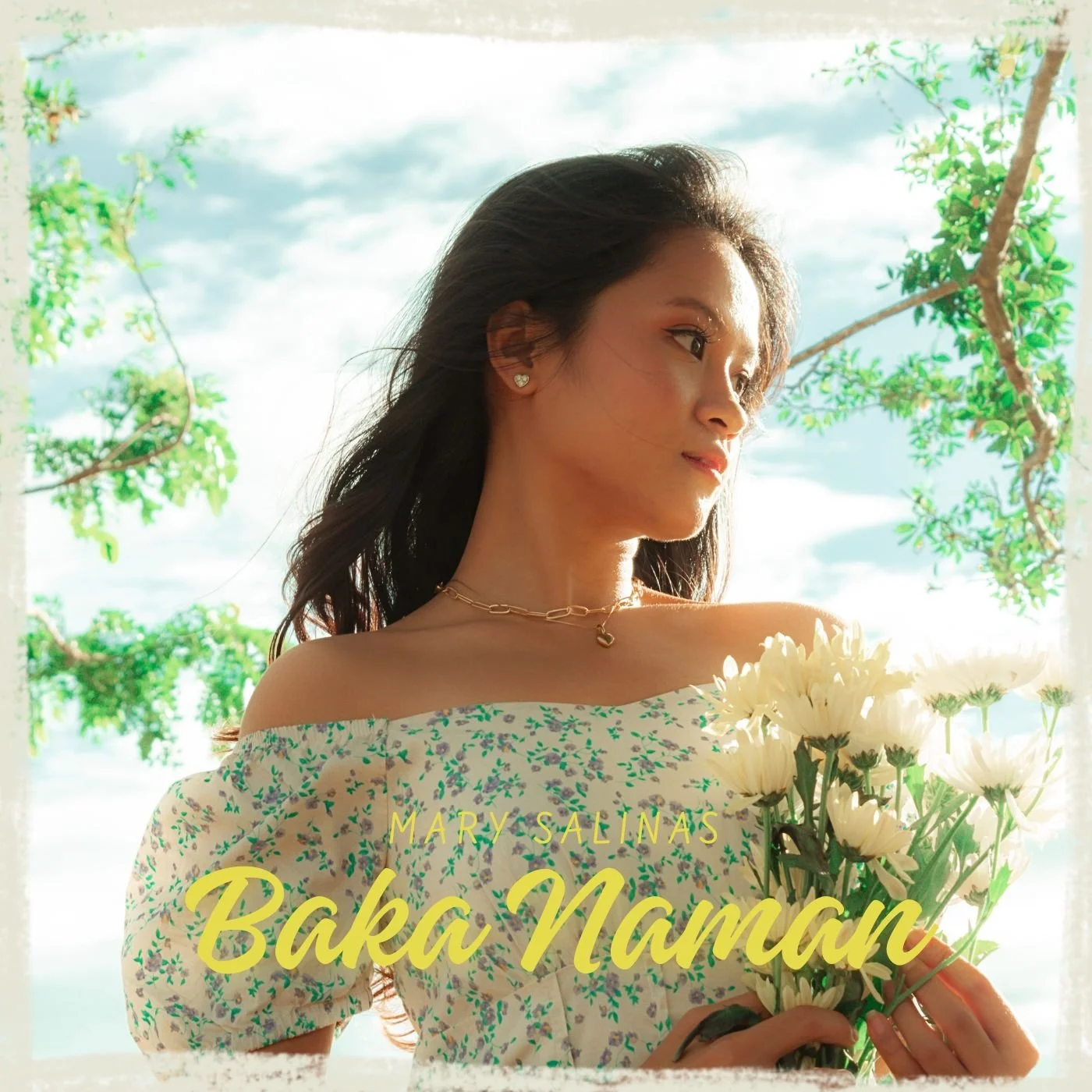 A woman with dark hair wearing a floral off-shoulder dress, smiling and holding white flowers, standing outdoors near green trees, with a partly cloudy sky in the background.